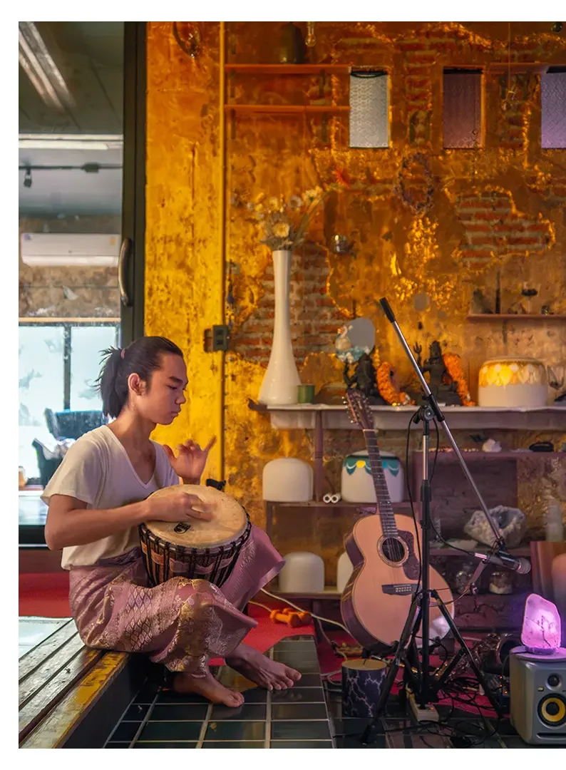 A young woman with dark hair tied back, sitting on a raised platform, playing a hand drum in a cozy, warmly lit room with a yellow brick wall. There is an acoustic guitar, microphone stand, and musical equipment nearby, decorated with shelves, vases,