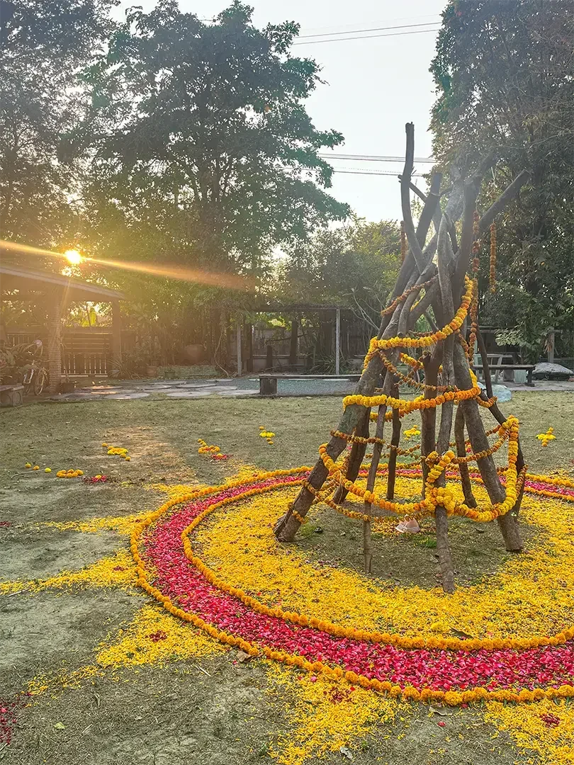 Colorful floral arrangement in the shape of a conical structure, decorated with yellow marigold garlands, set on a ground with pink and yellow flower petals forming a circular pattern around it, during sunset in an outdoor area.