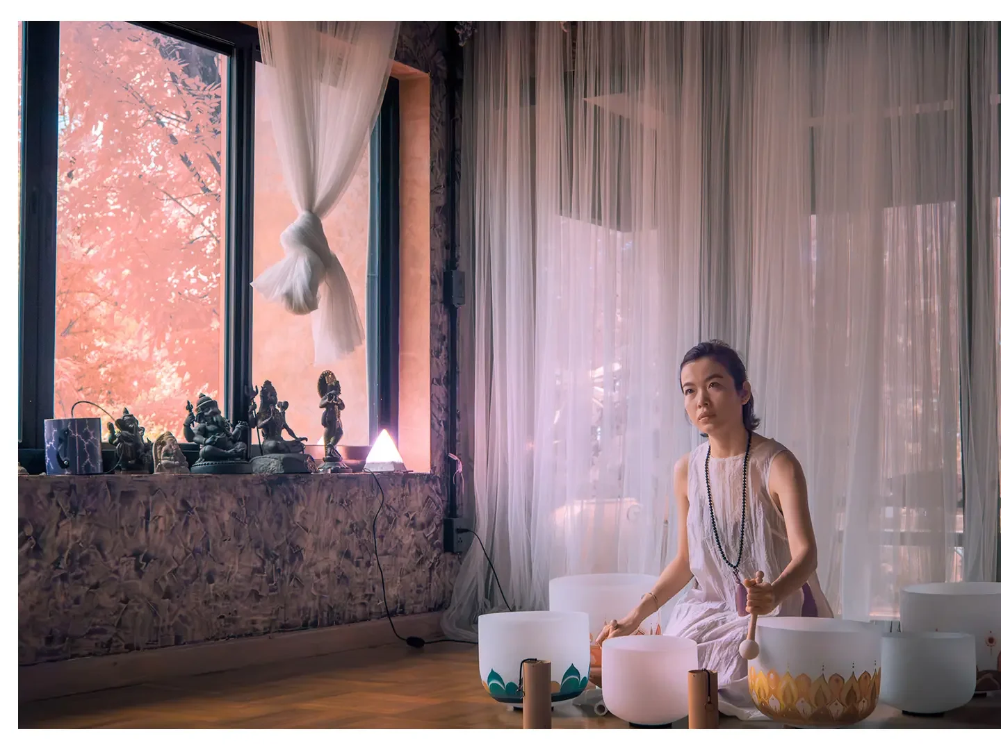A woman sitting on the floor in a serene room with sheer curtains, surrounded by singing bowls and statues of deities, with a large window showing pink autumn leaves outside.