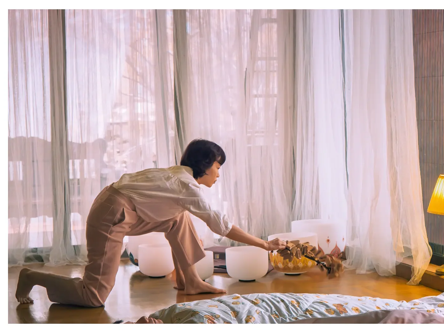 A woman on her knees, making percussion sound on the floor next to several white singing bowls, near a window with sheer curtains. There is a person lying on the bed nearby with part of their arm and hand visible.
