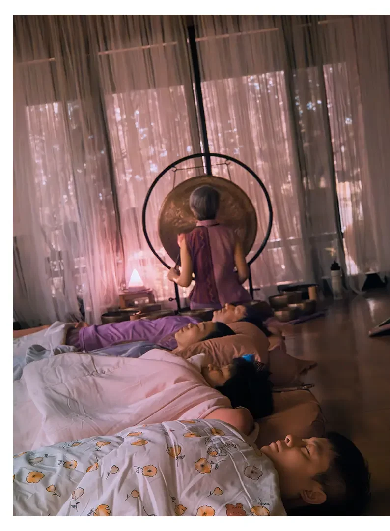 People lying down and sleeping on pillows, a woman sitting up in front of a large gong, with muted pink lighting coming through curtains in the background.