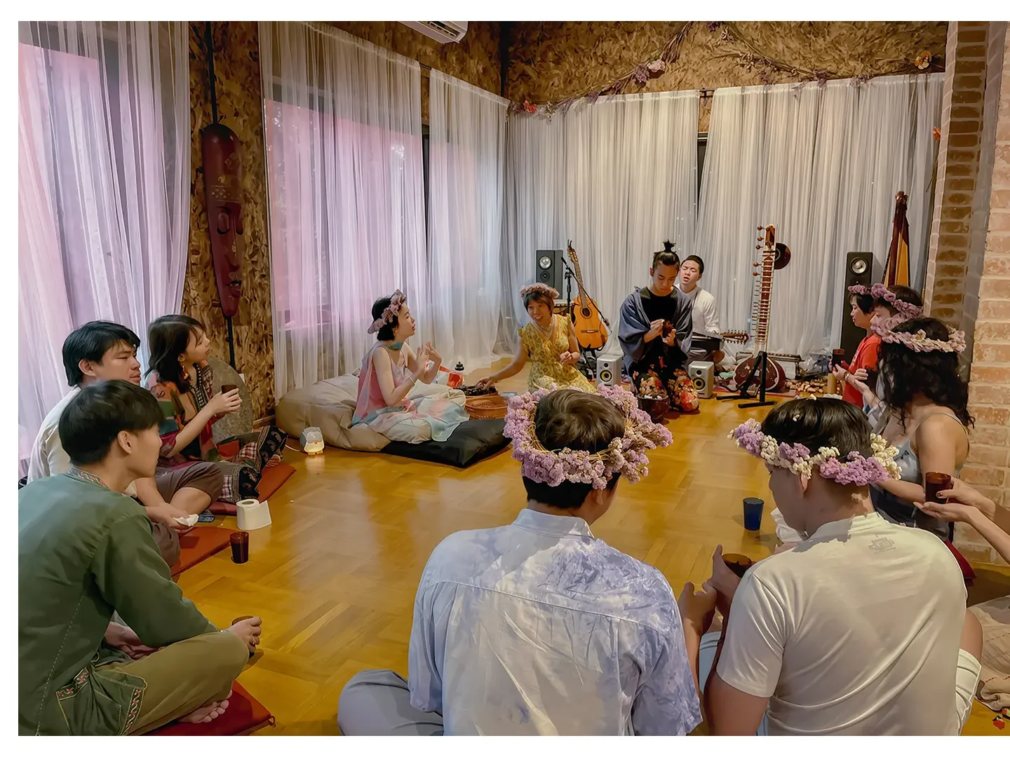People gathered in a cozy room with wooden flooring and sheer curtains, participating in a cultural or musical event. Some are sitting cross-legged on the floor, wearing flower crowns. There are musical instruments like guitars and traditional string