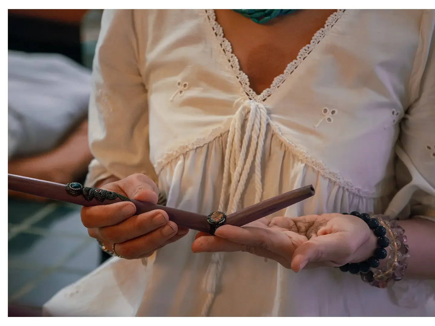 Close-up of a woman wearing a cream-colored blouse with lace details and tying a knot in a wooden stick with decorative accents. She has bracelets on her wrist and a ring on her finger.