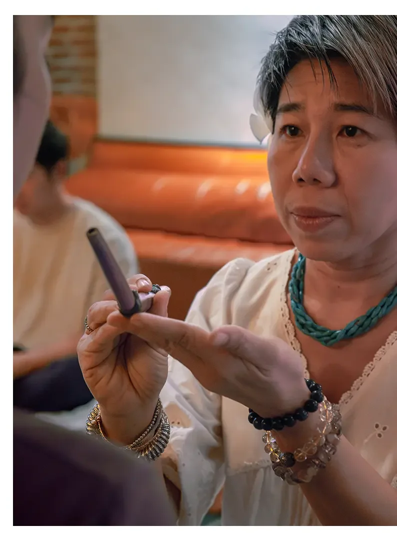 A woman with gray hair and multiple bracelets on her wrist, wearing a white blouse and turquoise beaded necklace, holding a rapeh tube and looking at another person.
