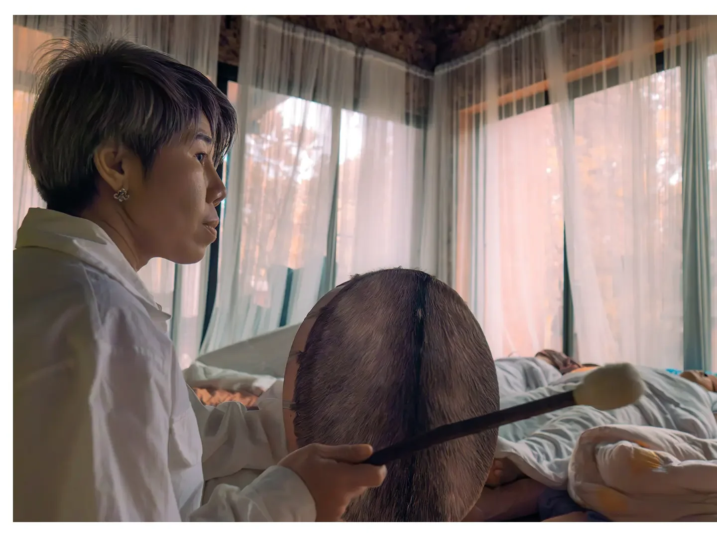 A woman with short hair and earrings is holding a shamanic drum in a room with large windows and curtains, with people lying on beds in the background.