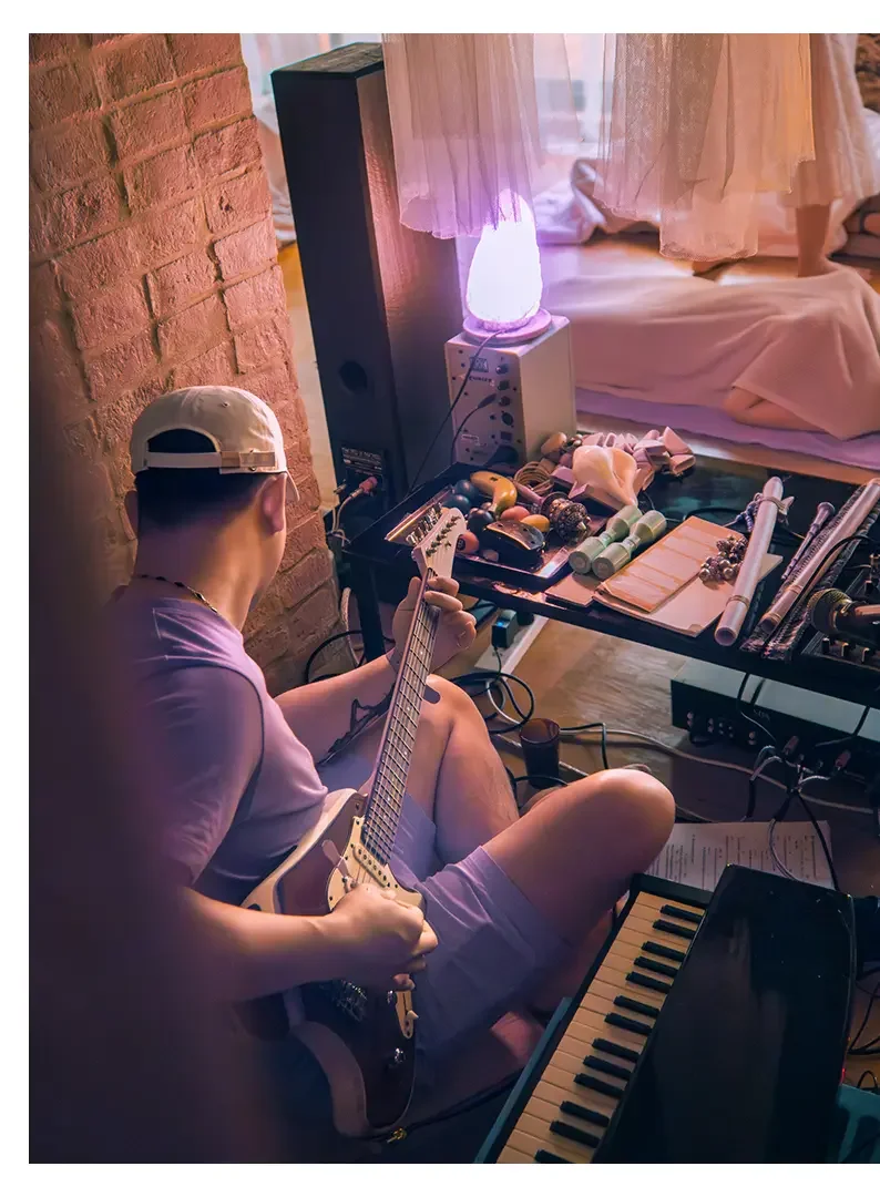 A person wearing a cap and shorts sitting on the floor playing an electric guitar in a cozy room with a brick wall, a bed with light-colored curtains, and a table filled with various objects including a small lamp, a keyboard, and other musical or de