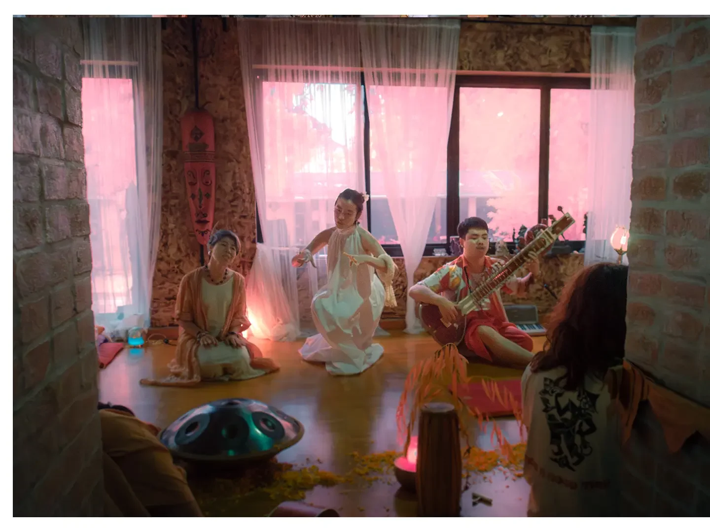 Indoor scene with three people performing. One woman sitting on the floor, another woman dancing, a man playing guitar. The room has pink curtains, stone walls, and wooden floors, with decorative items and flowers.