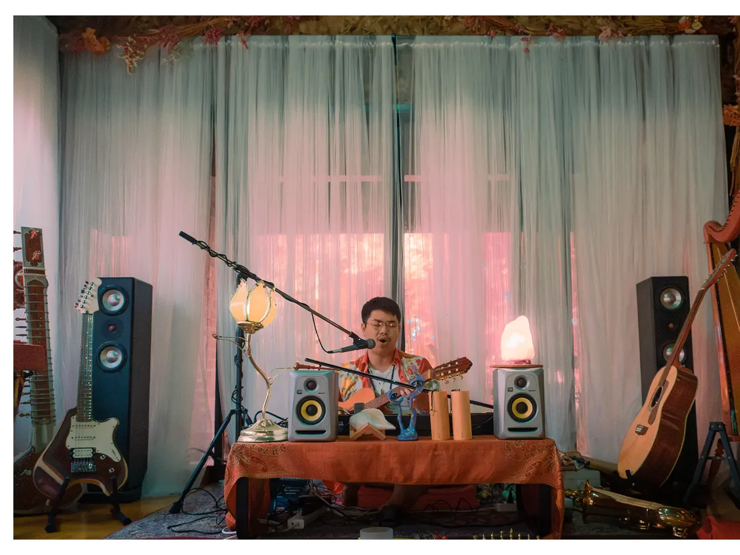 Man performing music in a cozy room with guitars, speakers, and warm lighting, with white curtains and wood decor.