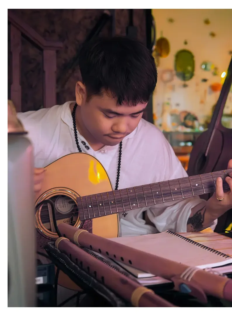 A young man playing an acoustic guitar with a notebook and flute in front of him, in a cozy, decorated space.