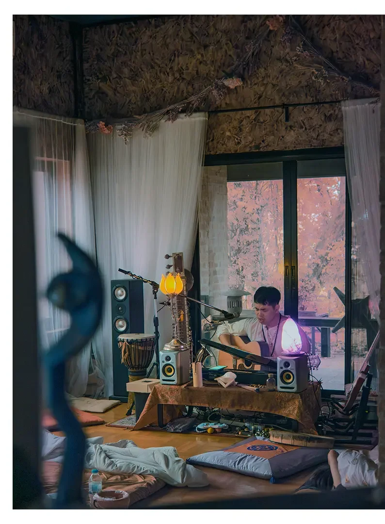 Young man playing an acoustic guitar in a cozy indoor space decorated with musical instruments, speakers, and a floral lamp, with a view of fall foliage through sliding glass doors.