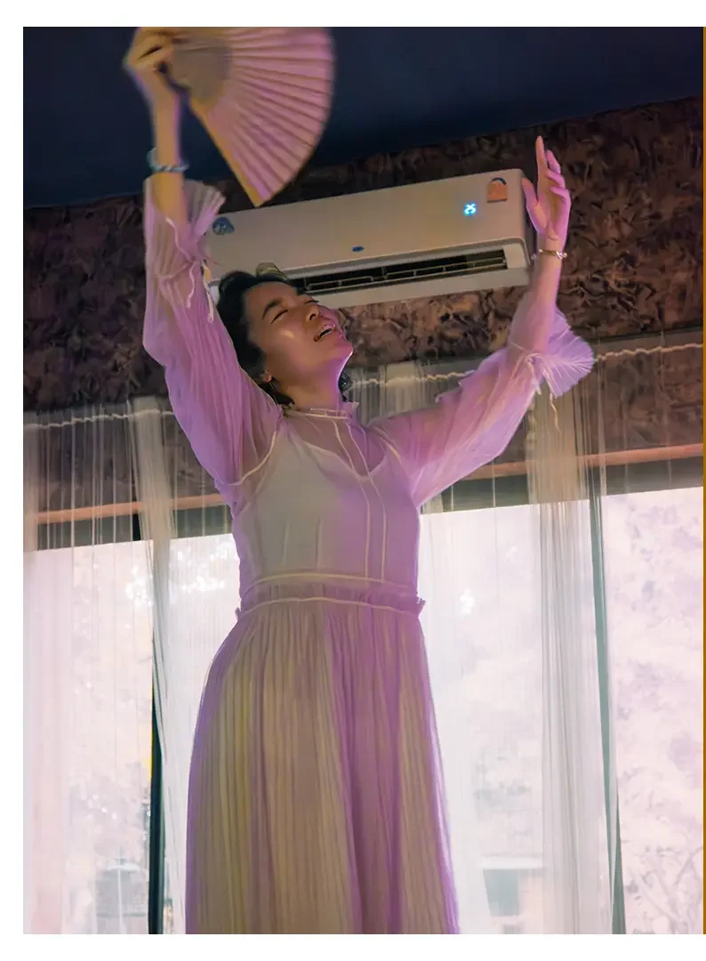 A woman smiling and joyfully dancing while holding a pink fan with her arms raised, standing in front of a window with sheer curtains.
