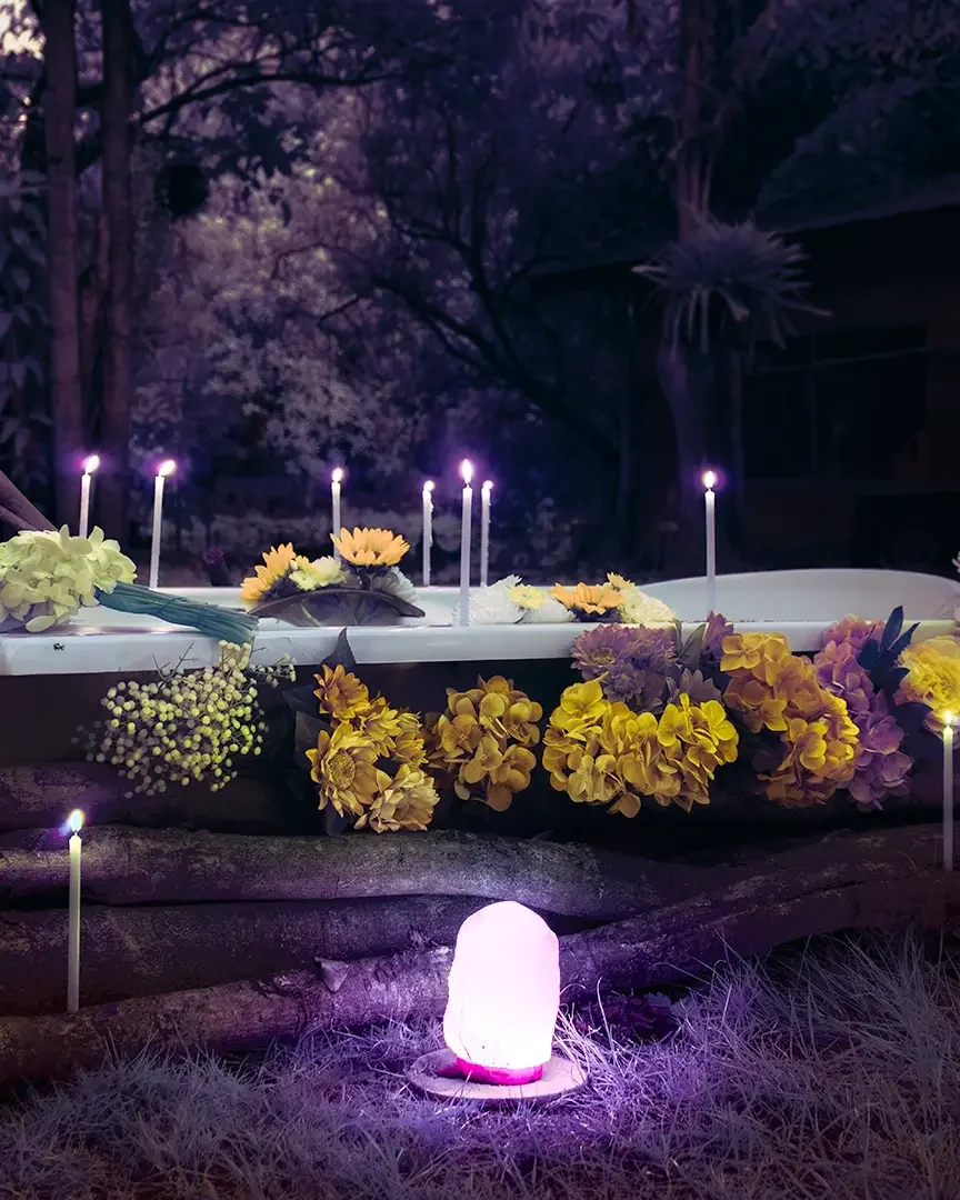 An outdoor scene with lit candles and flowers arranged around a bathtub and a glowing purple light in front, set against a dark, forested background.