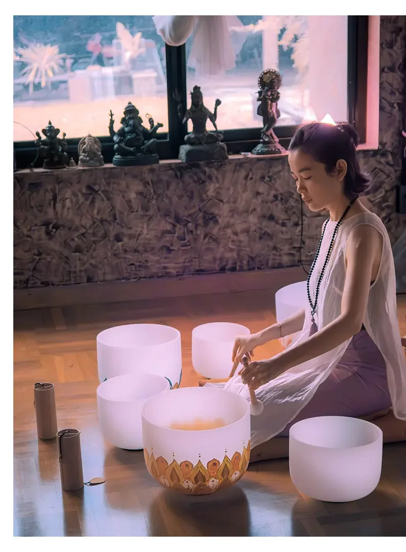 A young woman sitting on a wooden floor playing crystal singing bowls, with a window behind her displaying decorative statues and an outdoor scene.
