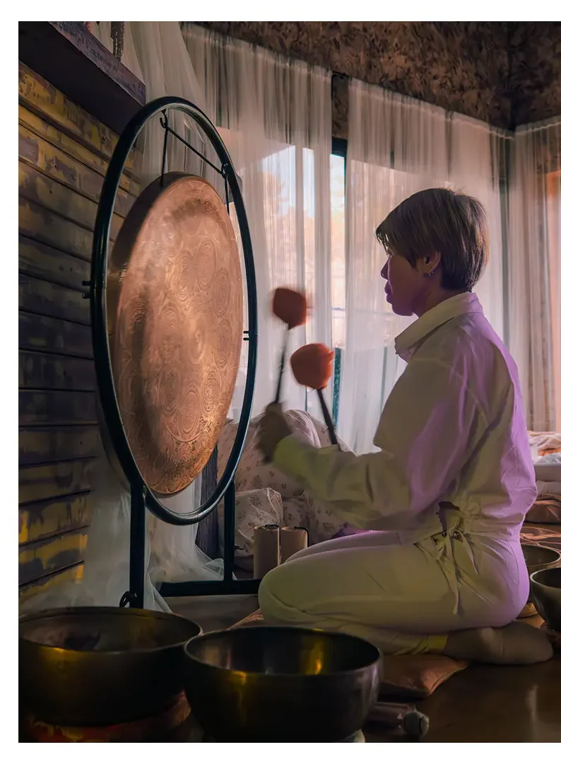 A woman in white clothing playing a large gong with mallets in a cozy room with wooden walls and sheer curtains, sunlight coming through the window.
