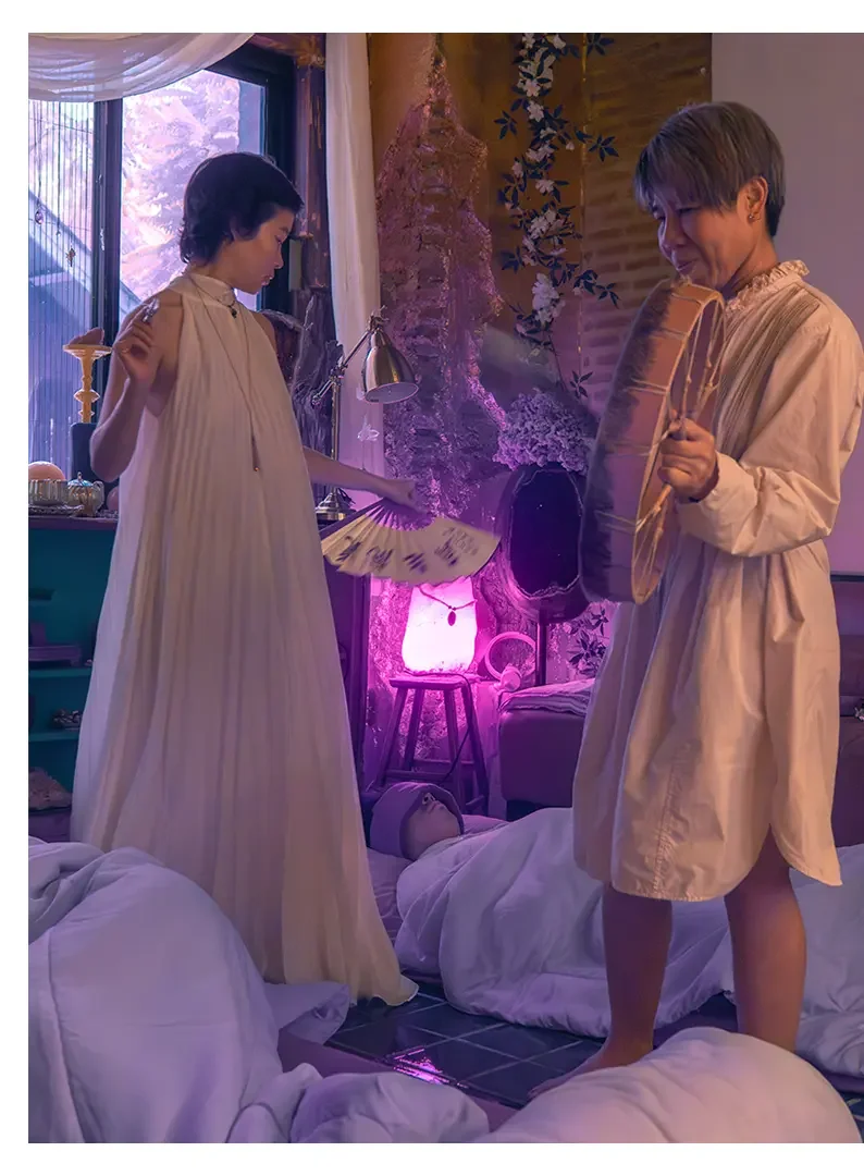 Two women dressed in beige robes stand in a softly lit room with a pink glow, some people lying on the floor covered with white sheets, and a person asleep in the background. One woman holds a fan, and the other holds a drum, with a purple-lit lamp, 
