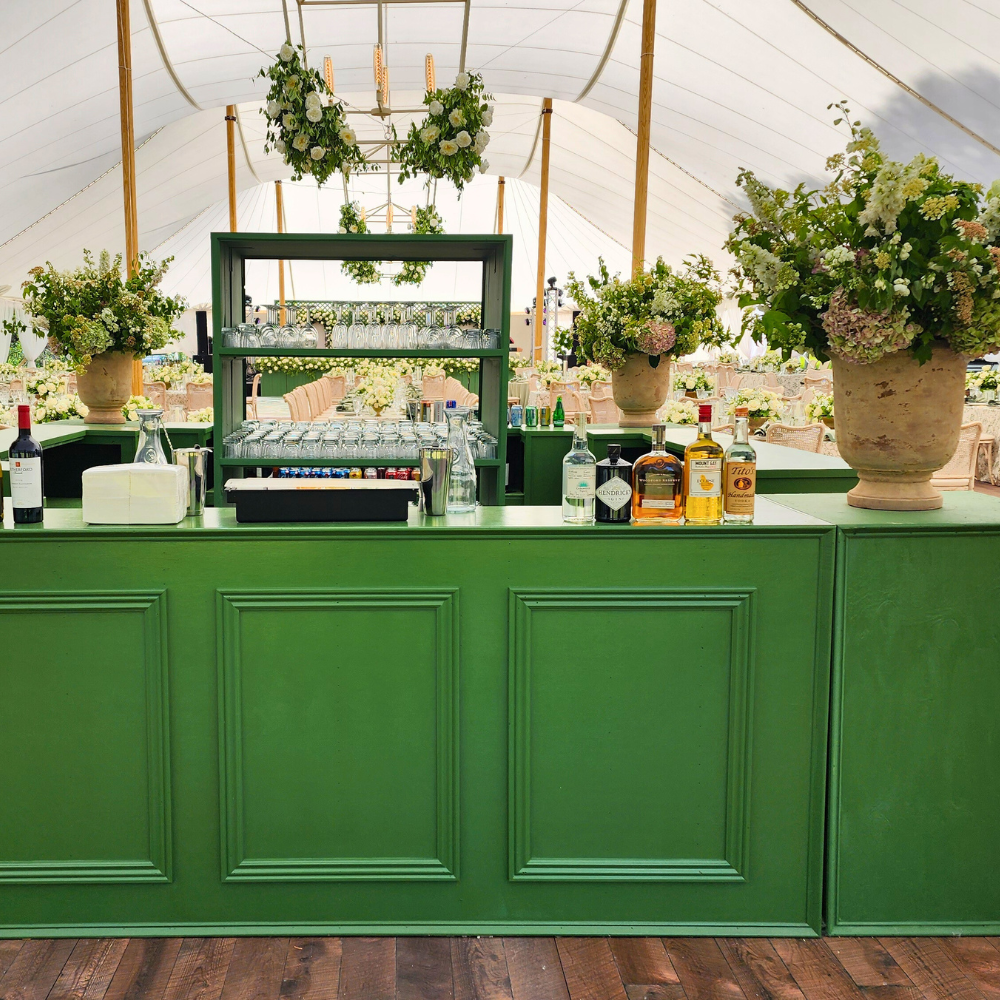 A green bar setup at a wedding venue with large floral arrangements, bottles of liquor, and glassware inside a white tent.