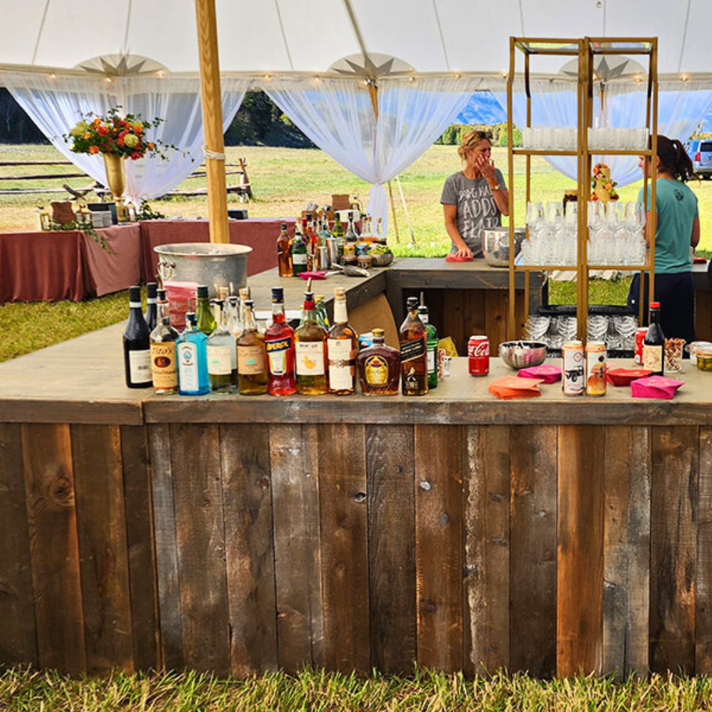 Bar setup inside a white event tent with a table of various alcohol bottles in front, and two women preparing drinks in the background. The tent is outdoors in a grassy area with mountains in the distance.
