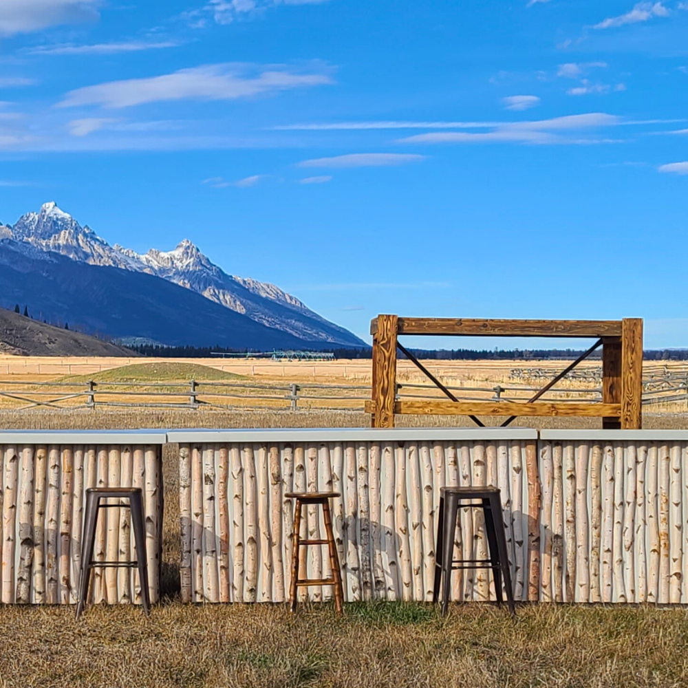 Seaside outdoor bar with wooden stools, a beach fence, and mountain range in the background under a blue sky with clouds.