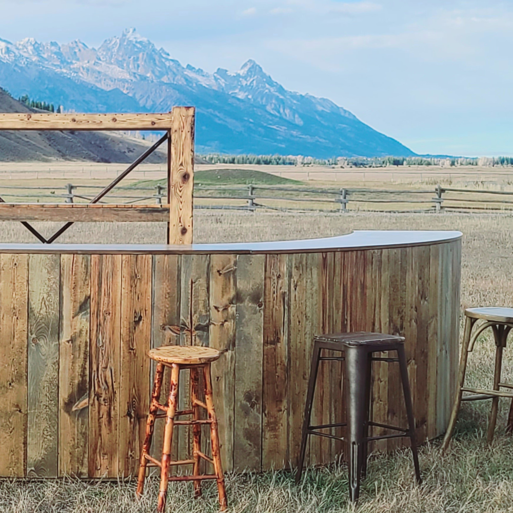 An outdoor bar made of wood with three bar stools, set against a mountain landscape with grass and a wooden fence in the background.