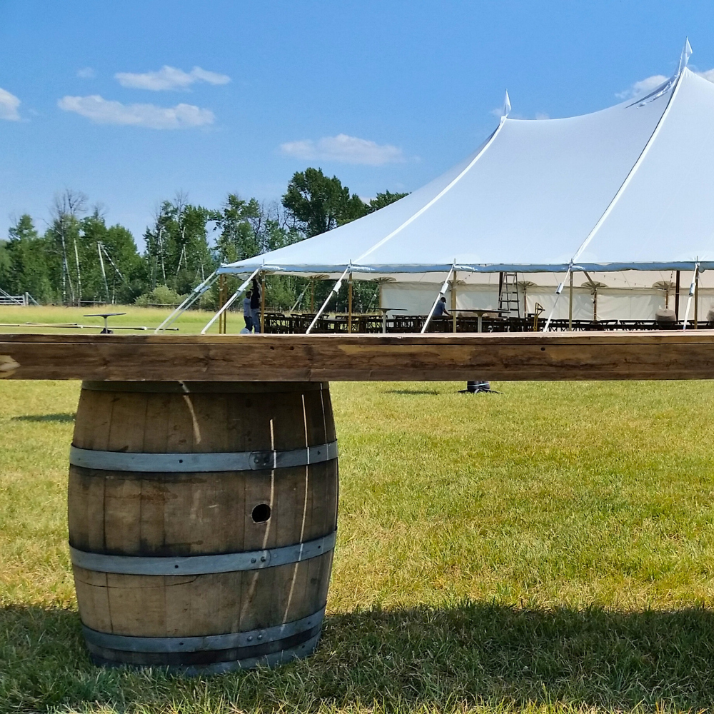 Outdoor event setup with a white tent, tables, a ladder, and people working on the tent frame on a grassy field under a blue sky.