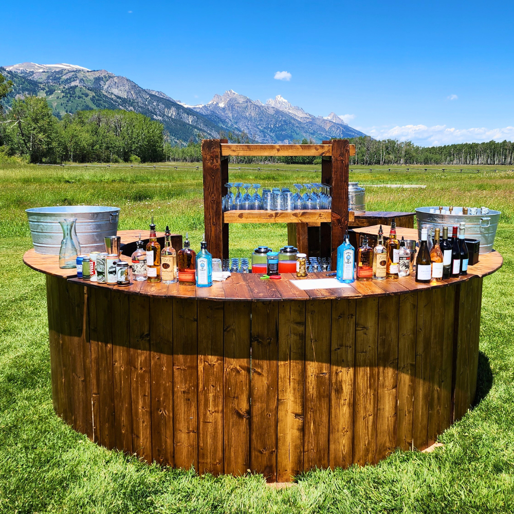 Outdoor bar setup with various bottles and glasses in a field, mountains in the background on a sunny day.