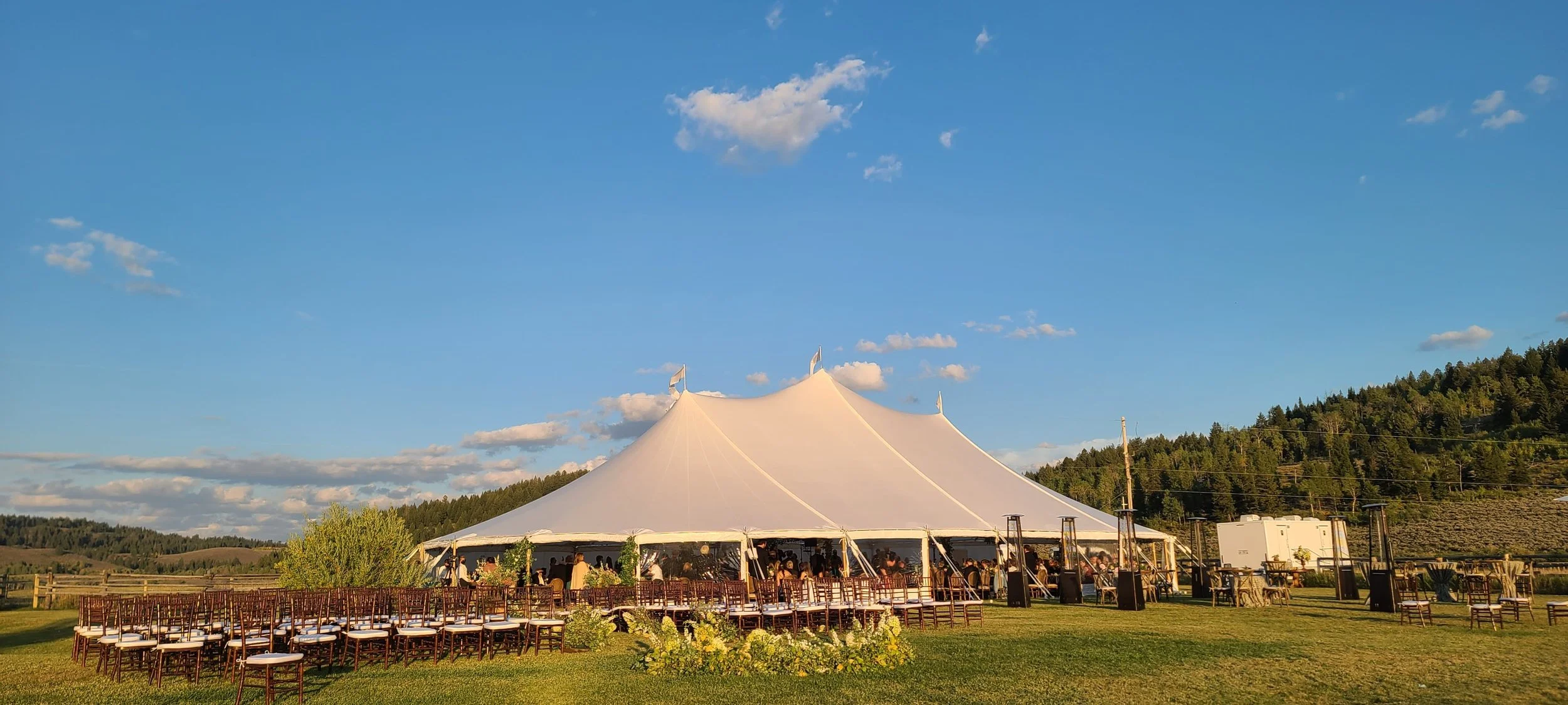 An outdoor event tent was set up in a grassy field in Jackson, Wyoming, with chairs arranged for a gathering, surrounded by rolling hills and trees under a partly cloudy sky.