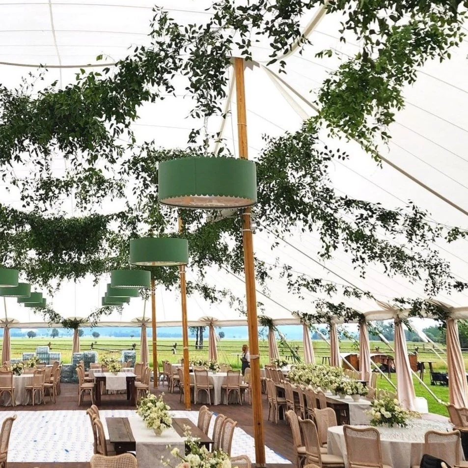 Inside a decorated event tent with round tables, floral centerpieces, and green hanging lamps, overlooking a field with a few people in the distance.