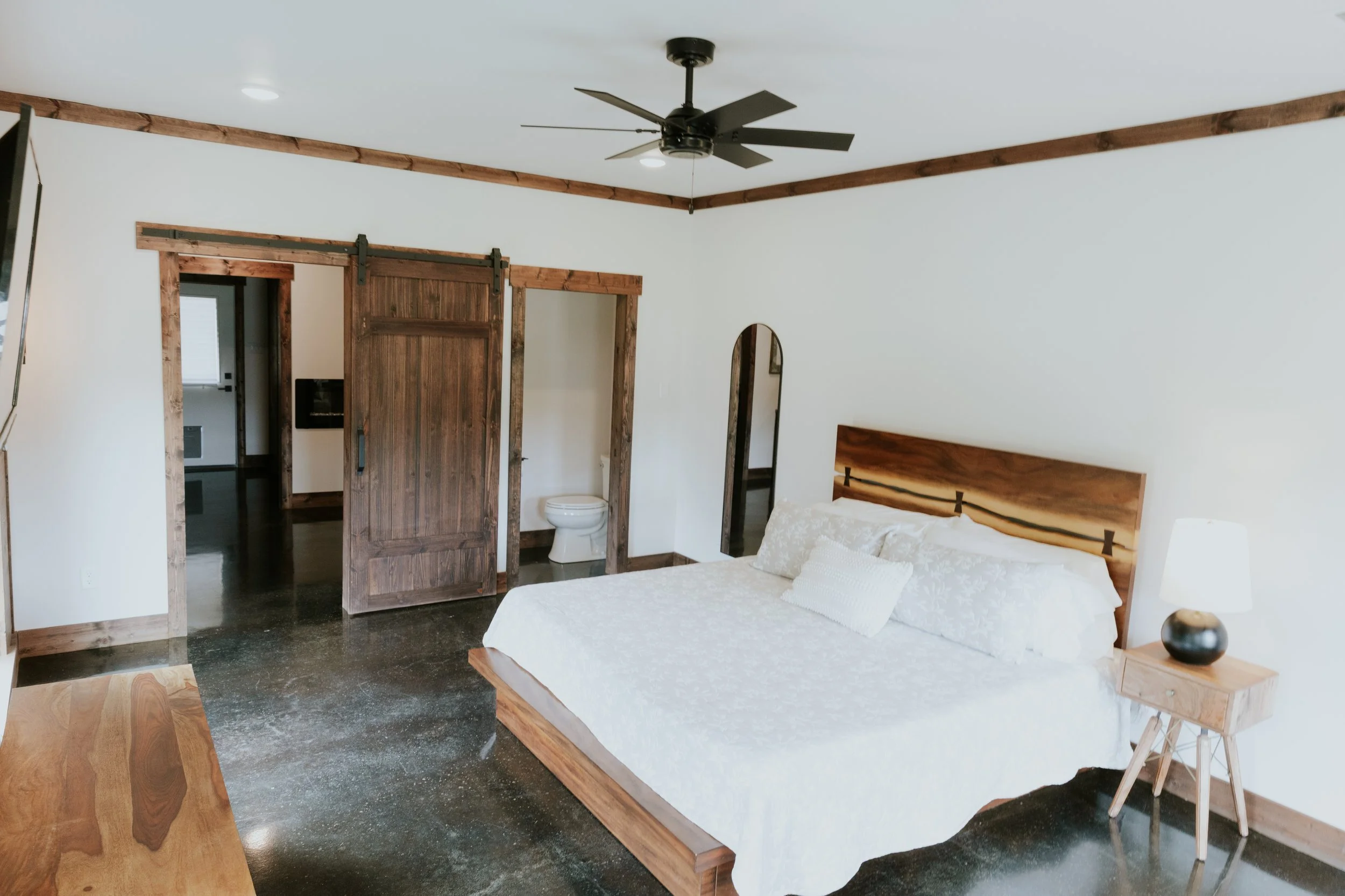 A bedroom with a wooden bed frame, white bedding, a wooden nightstand with a lamp, a ceiling fan, and a sliding barn door leading to a bathroom.