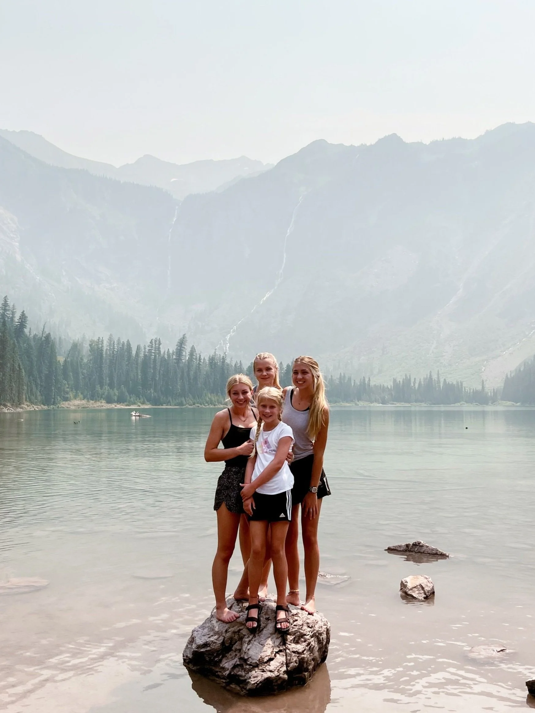 Four women standing on a rock in a lake with mountains and forests in the background.