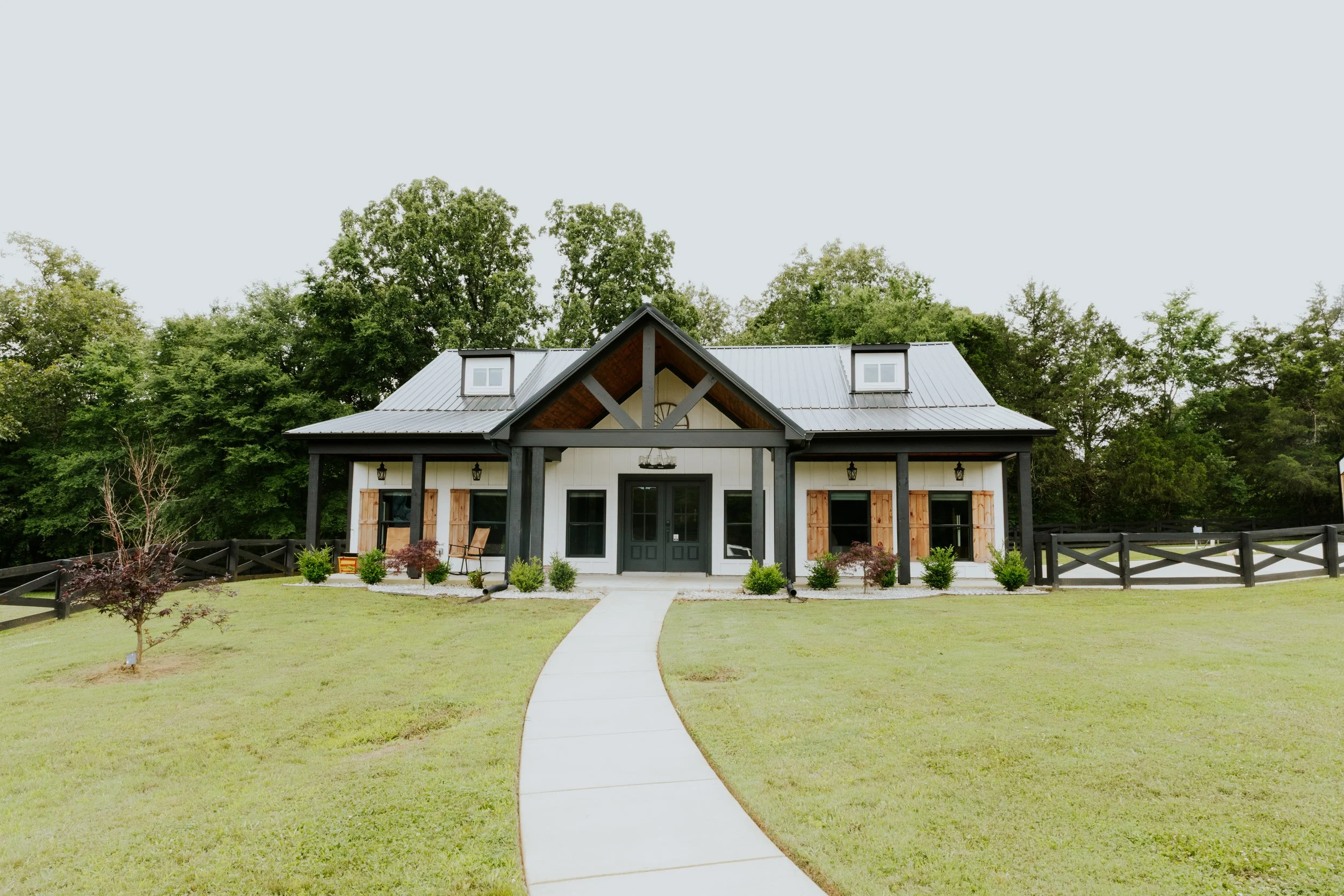 Front view of a modern farmhouse with a black and white exterior, wooden shutters, and a metal roof, surrounded by a green lawn and trees.