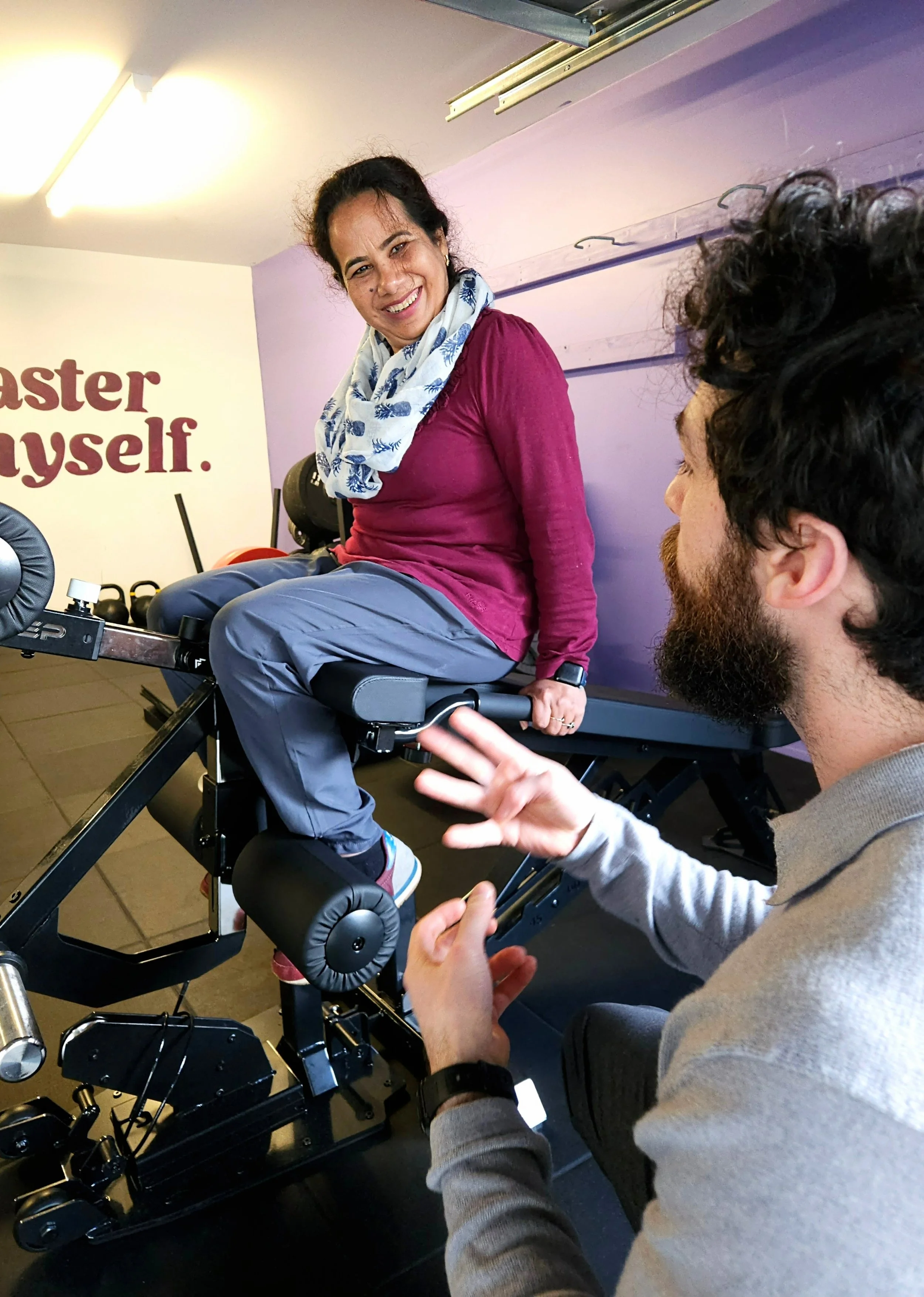 A local Karori woman using REP Leg Extension & Leg Curl Bench Attachment, smiling and engaging in conversation with Julian, founder of Craft Fitness in a gym setting with kettlebells in the background and a motivational sign on the wall that reads 'M