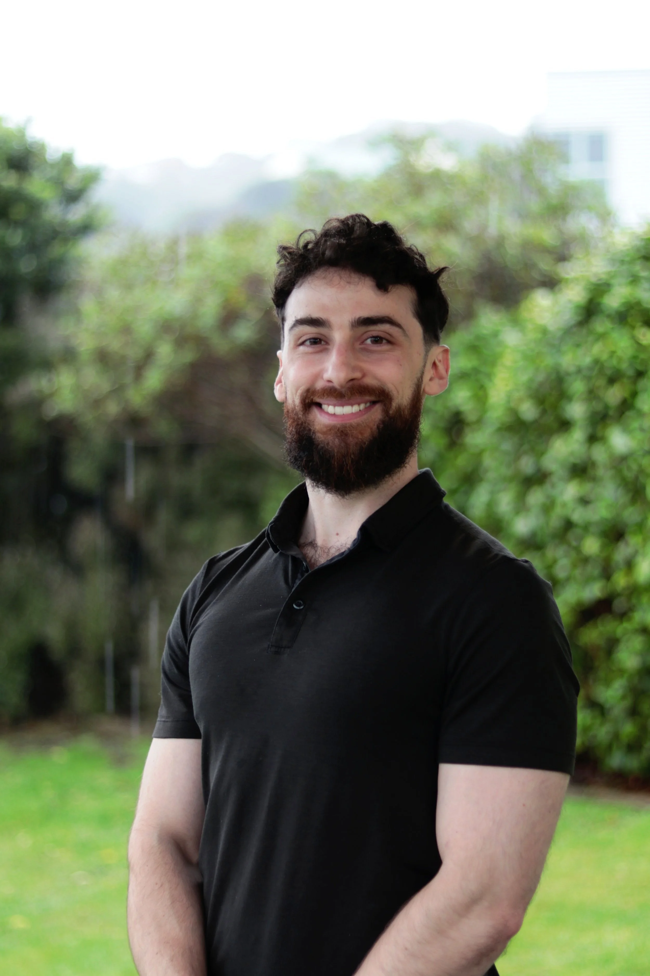 A photo of the personal trainer, who is a young man with dark curly hair, a full beard, and light skin smiling outdoors in front of green trees and a building in Karori