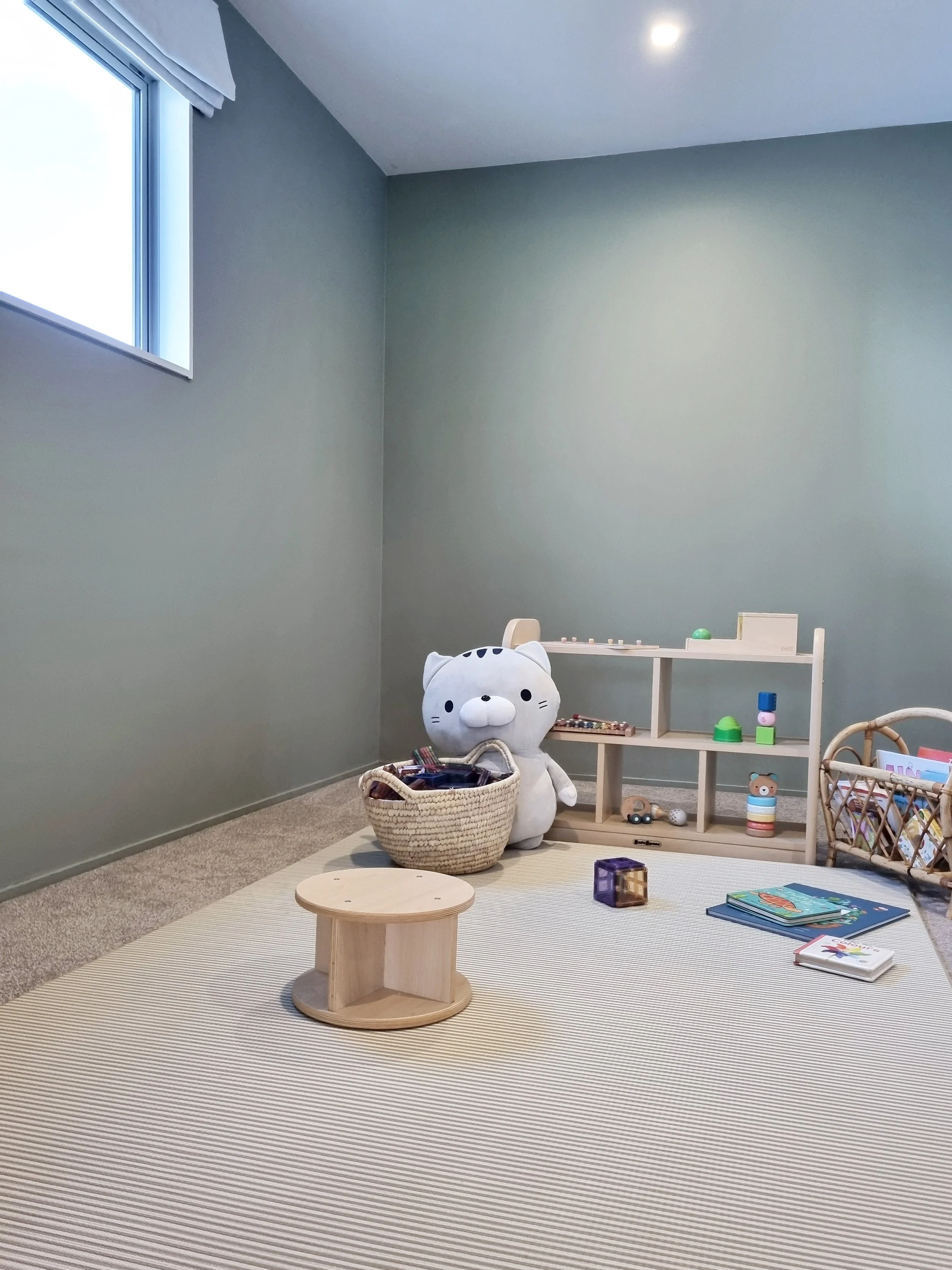 Children's playroom with a stuffed animal, wooden shelves, toys, books, and a small table on a striped rug.