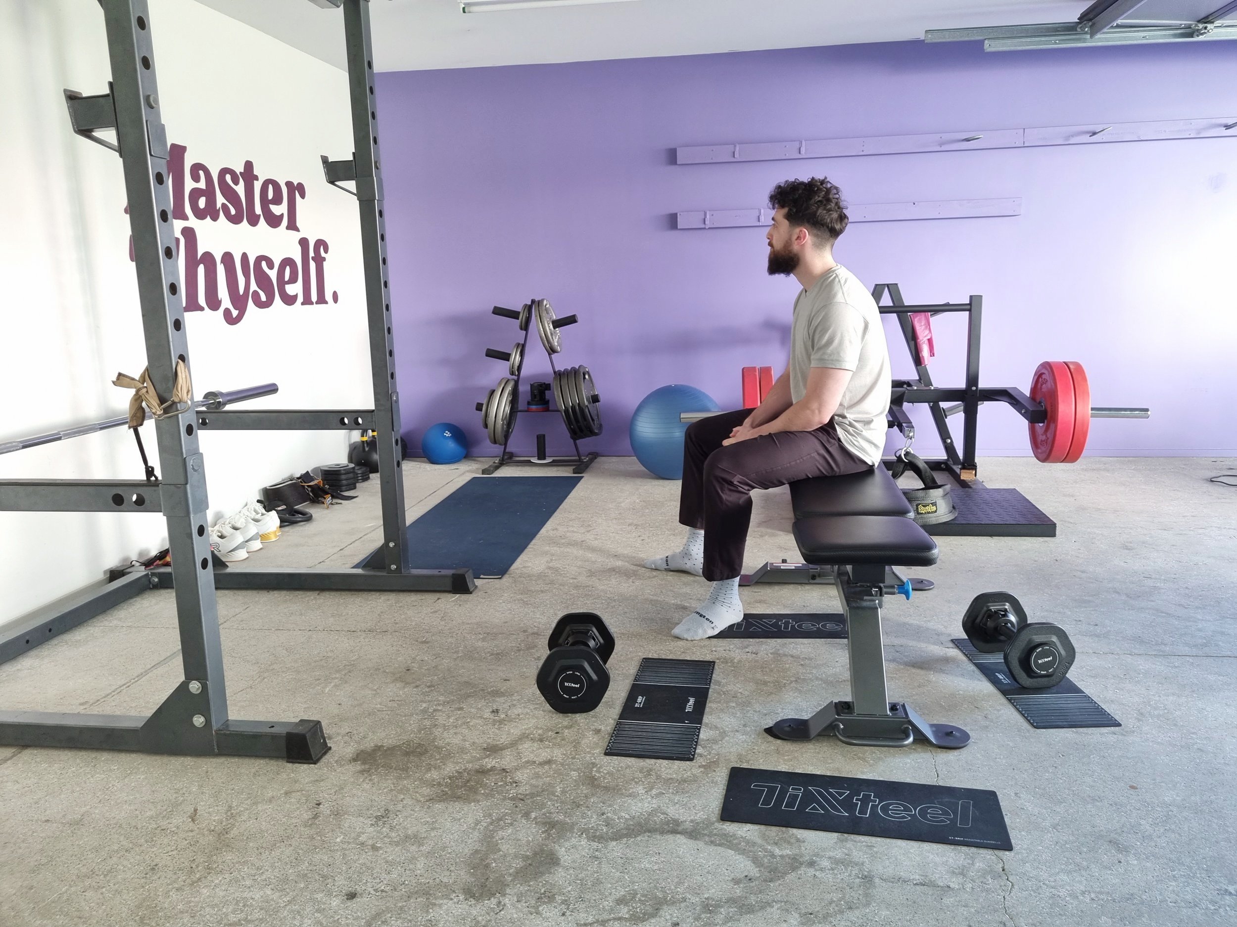 A man sitting on an adjustable workout bench with exercise equipment around him in a gym, with the words 'Master Thyself' painted on the wall.
