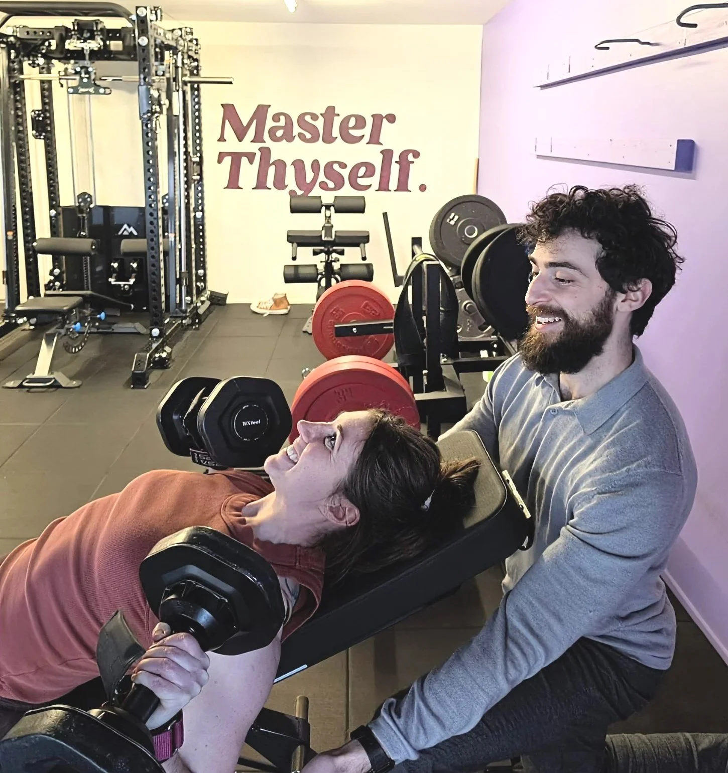 A local Karori woman doing a dumbell bench press, smiling, while being trained by Julian Etchegoin, founder and personal trainer at Craft Fitness Studio. The wall in the background has a motivational quote that says "Master Thyself." The gym contains