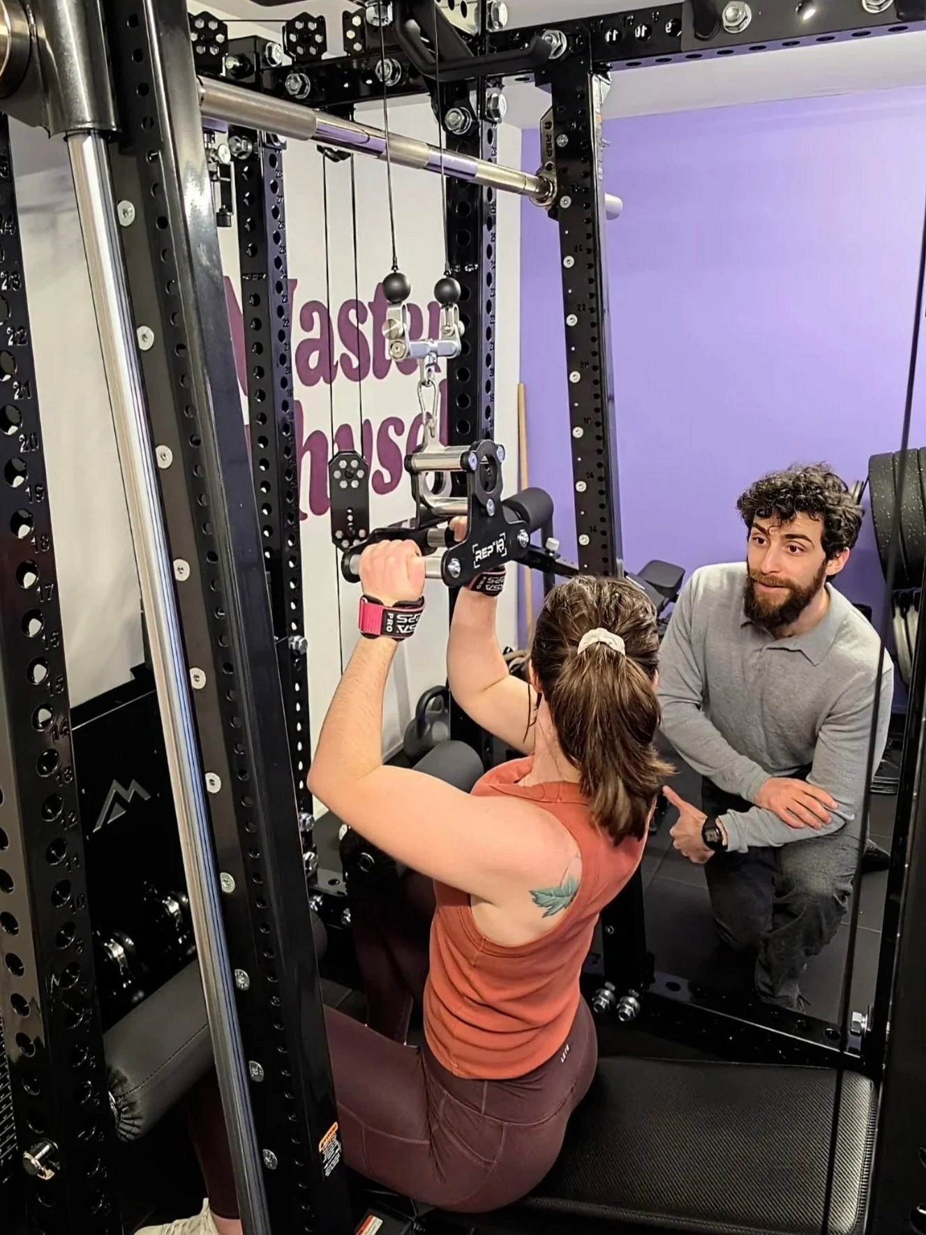A local Karori woman mid way through a lateral pull down exercise using the REP Ares™ 2.0 cable attachment. Trained by Julian Etchegoin, Craft Fitness Studio Founder and personal trainer. The gym has purple walls and various workout equipment.