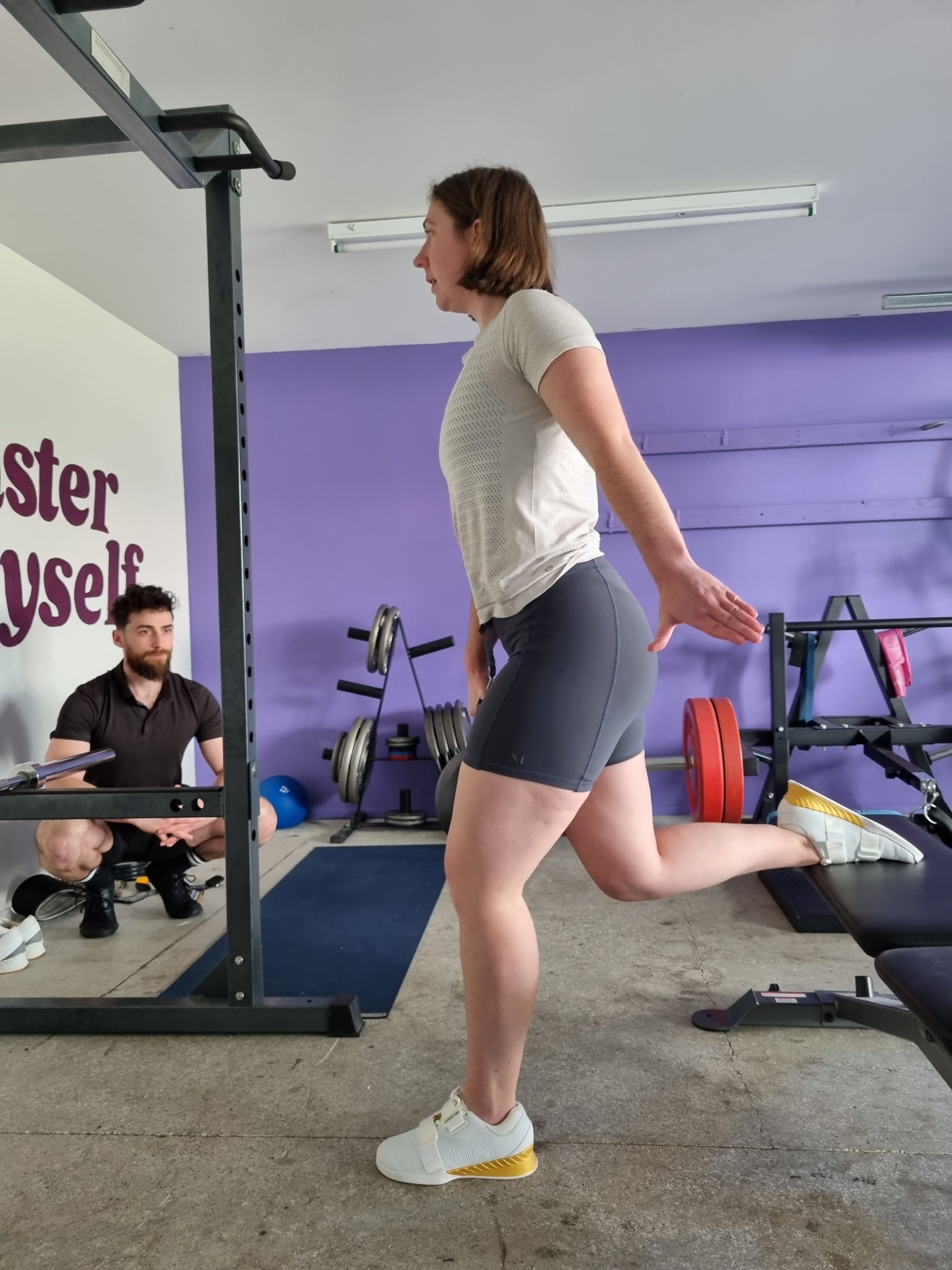 A woman performing a glute bridge exercise on a bench in a gym, with a man observing nearby.