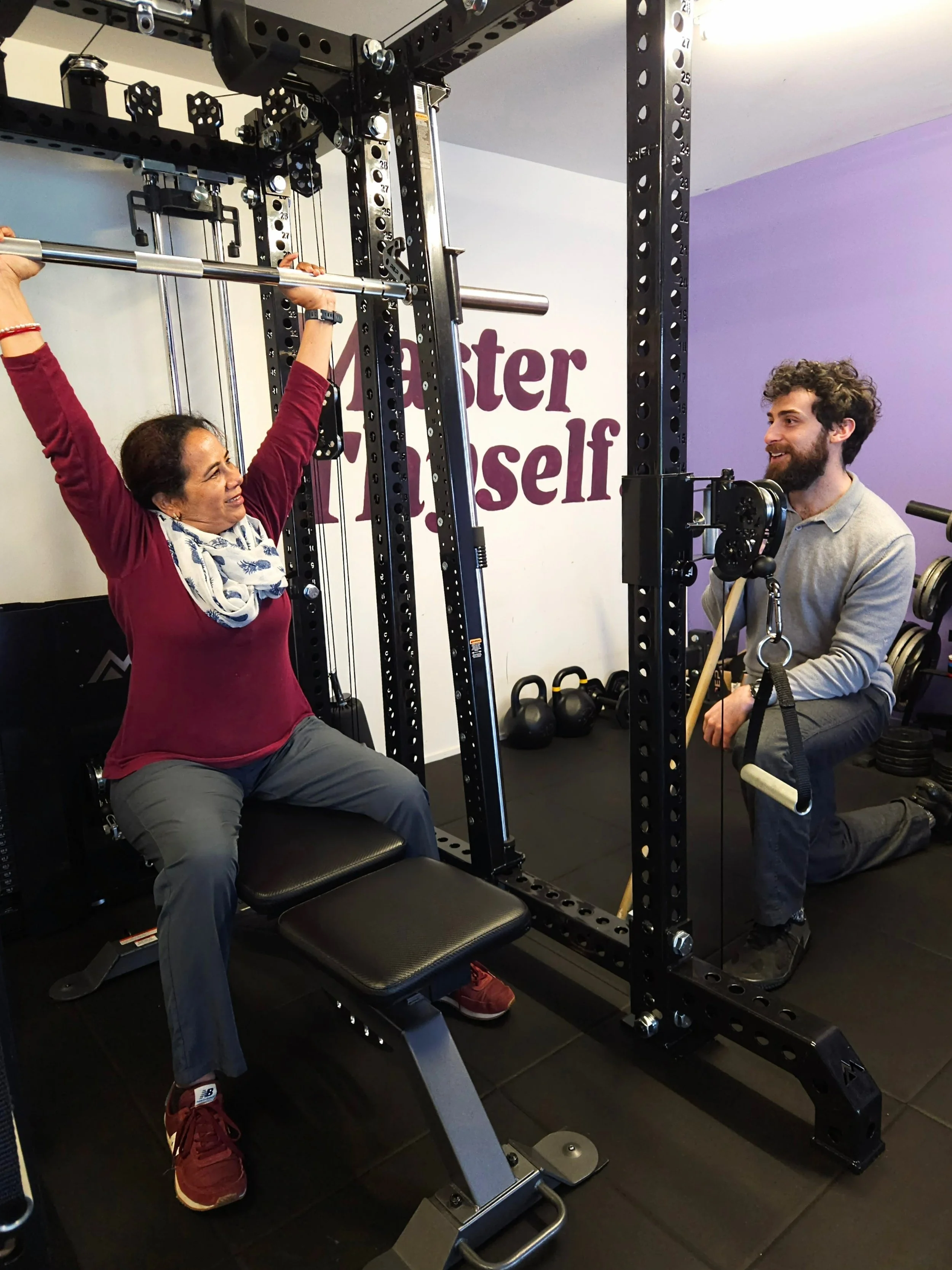 A local Karori woman being instructed through an overhead press exercise using the REP Smith Machine Rack Attachment. while her trainer Julian kneels beside her, monitoring her form. The gym has various weights and equipment around.