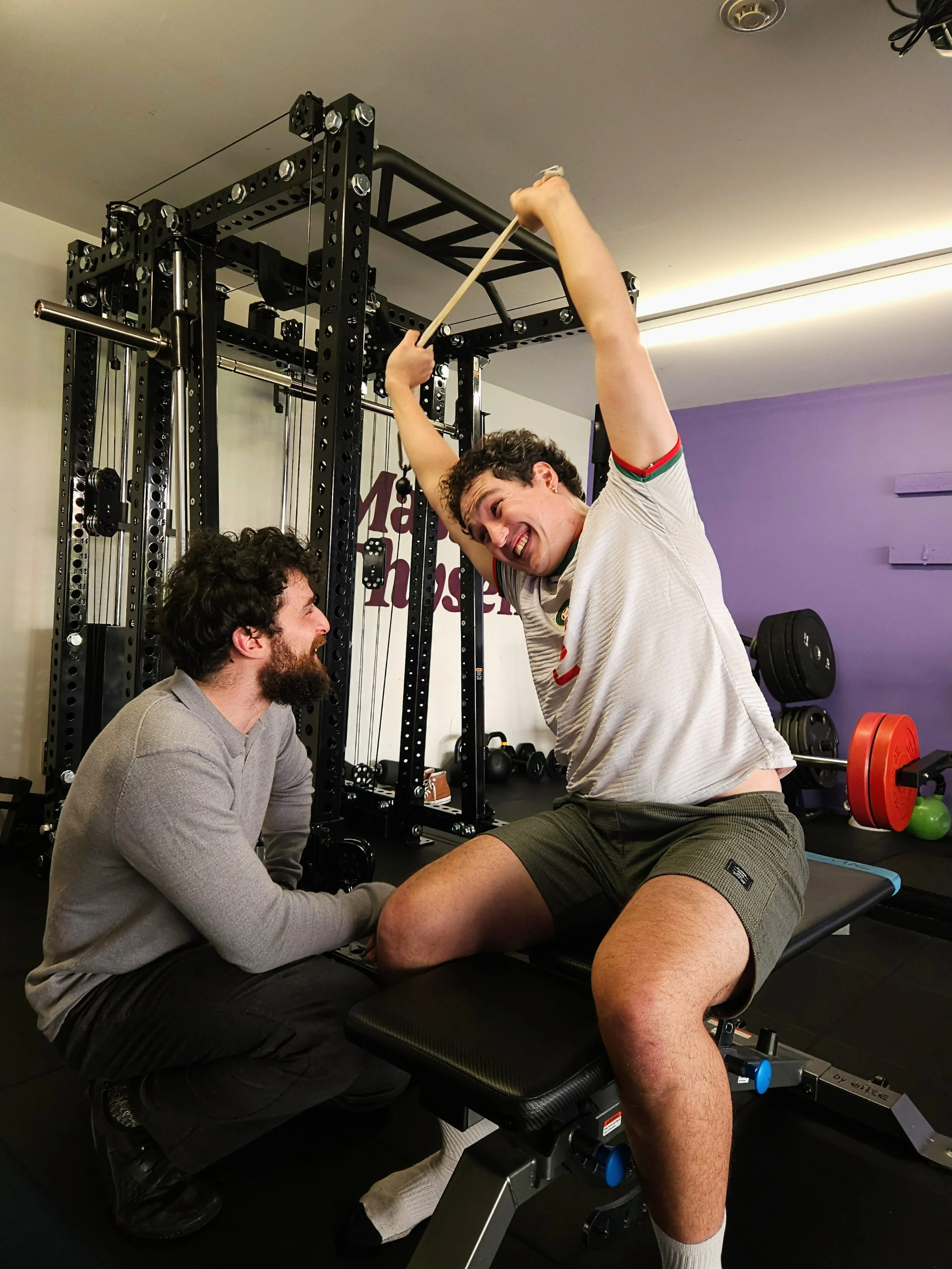 A man in athletic clothing doing a seated overhead cable side bend with the REP Ares™ 2.0 cable attachment. Trained by Julian Etchegoin, Craft Fitness Studio Founder and personal trainer. The gym has purple walls and various workout equipment.