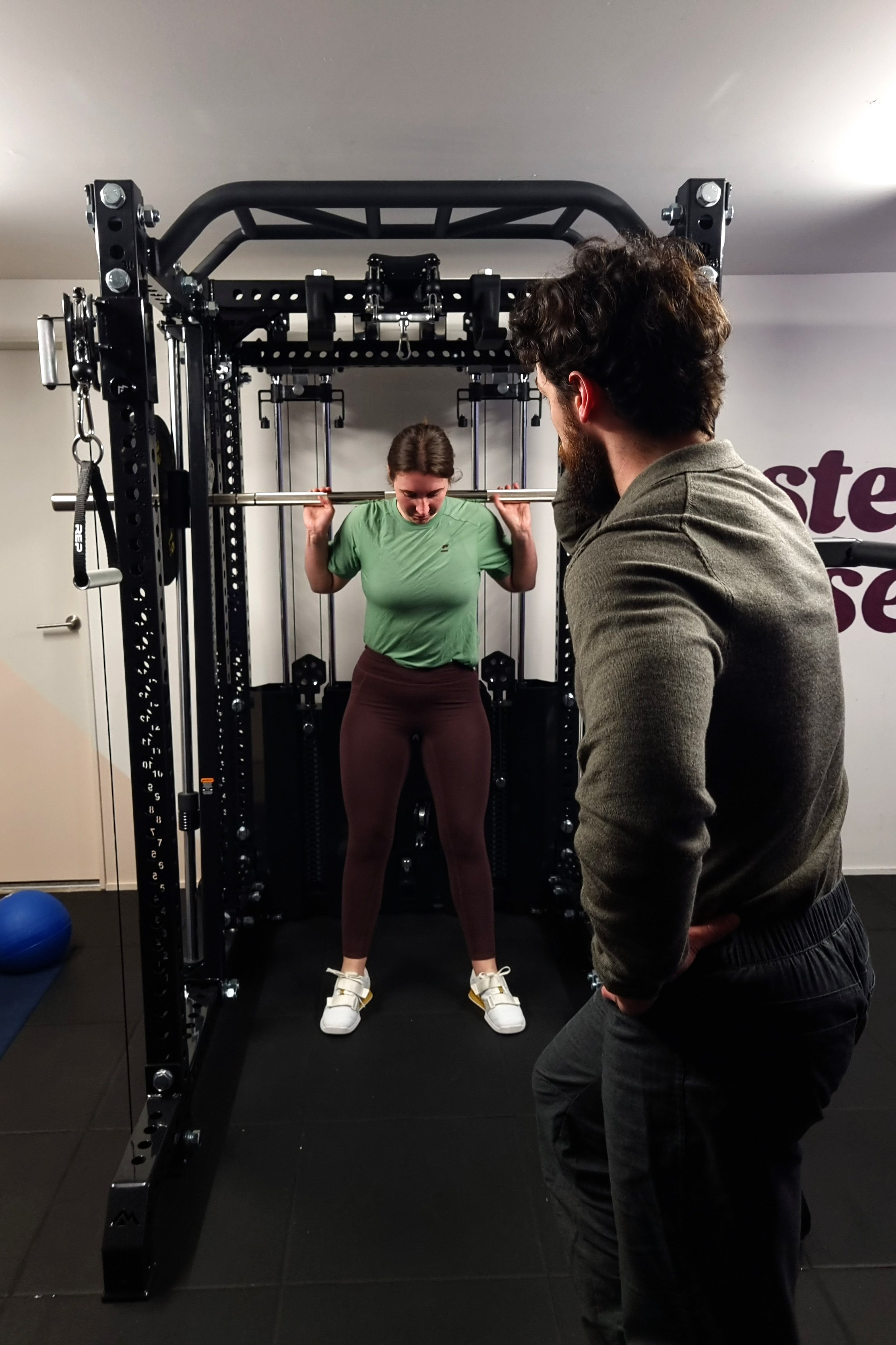 A woman is performing a squat using the REP Ares™ 2.0 Smith Machine Rack Attachment. Julian Etchegoin, Craft Fitness Studio Founder and personal trainer obverses form.