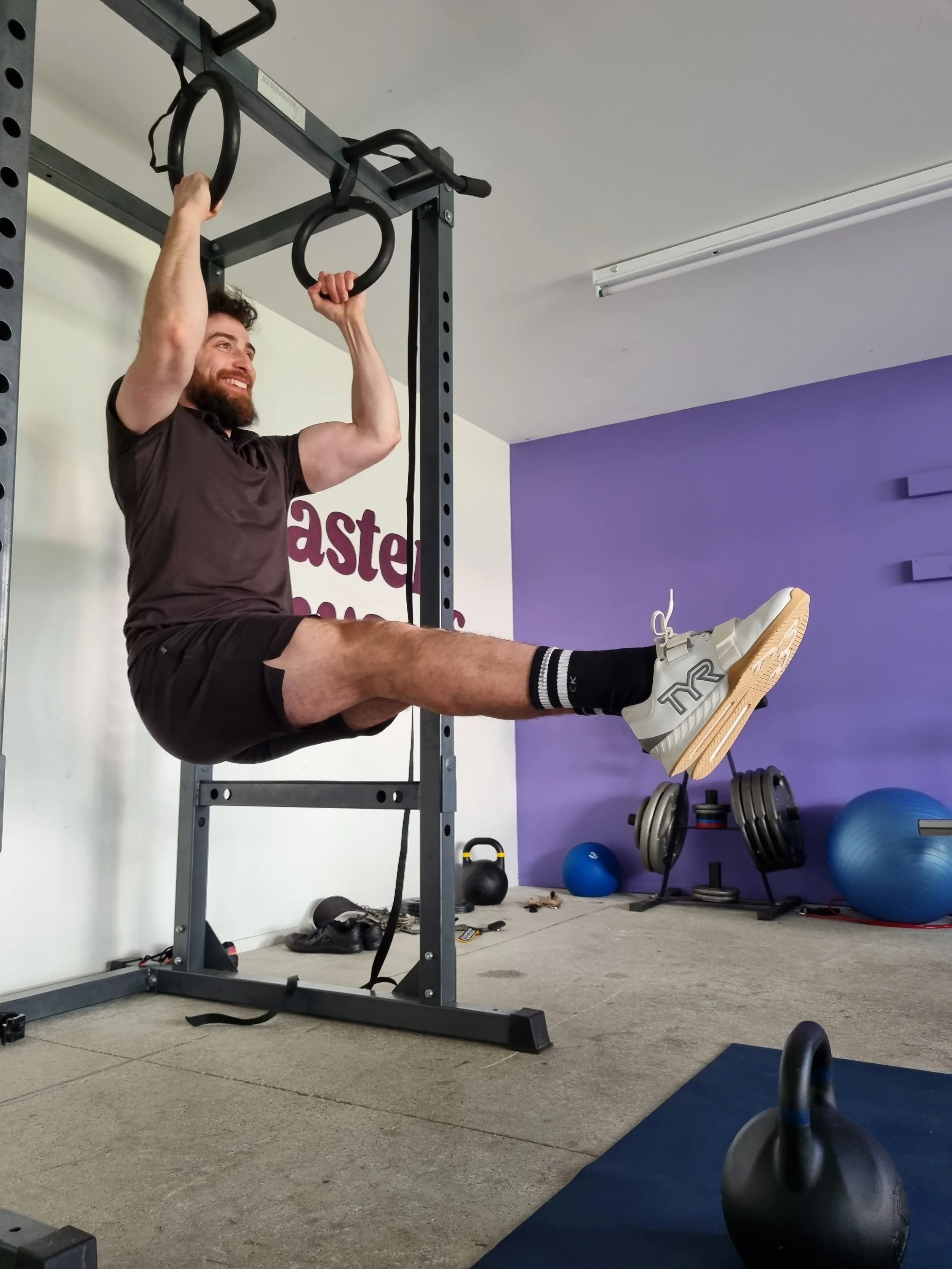 A man with a beard wearing a brown T-shirt, black shorts, and white sneakers is hanging from gymnastic rings attached to a black metal frame, with his legs extended straight out. He is smiling and appears to be exercising in a gym with purple and whi