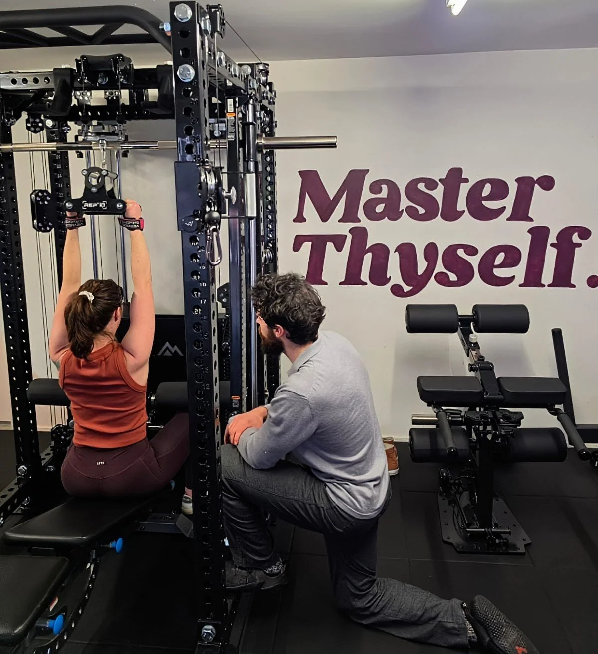 A woman performing a lateral pulldown exercise while being coached by her trainer Julian in his Karori personal training Studio. The wall behind them has the words "Master Thyself" written on it.