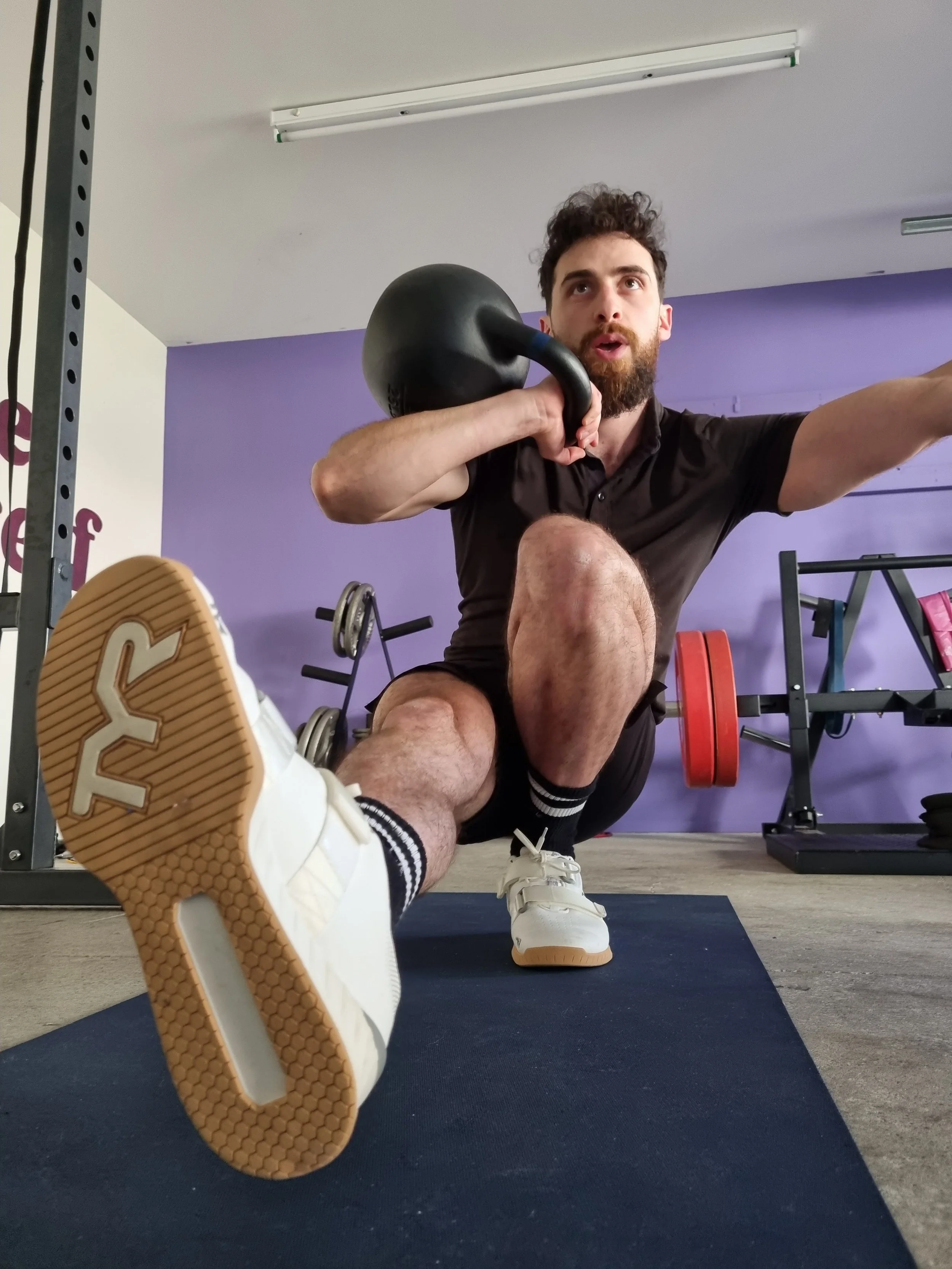 Man performing a kettlebell exercise in a gym, wearing black shorts, white sneakers with black socks, and holding a black kettlebell to his shoulder, with a purple wall and fitness equipment in the background.