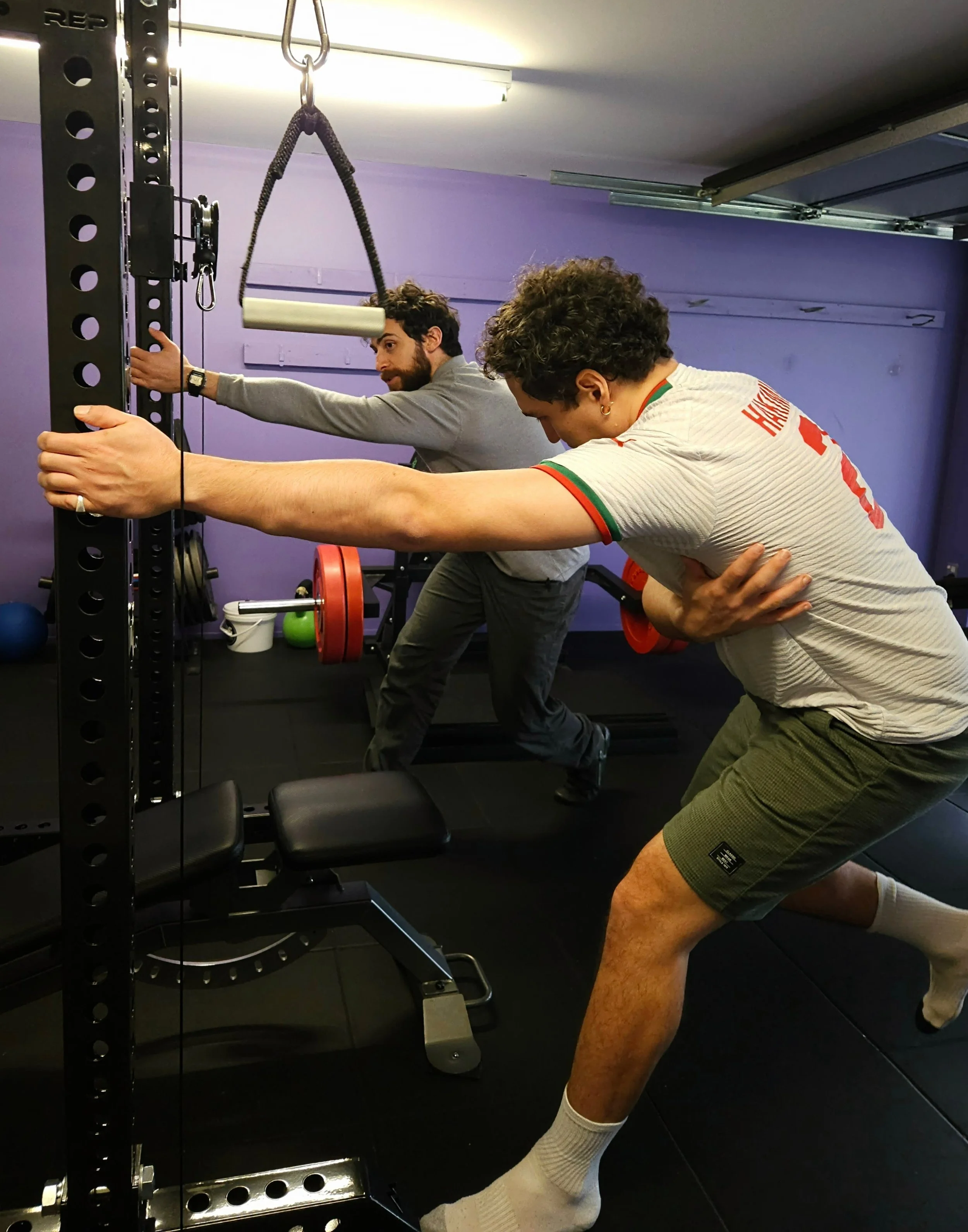 Two men in a gym stretching in front of a weightlifting rack with weights, resistance bands, and gym equipment in the background.