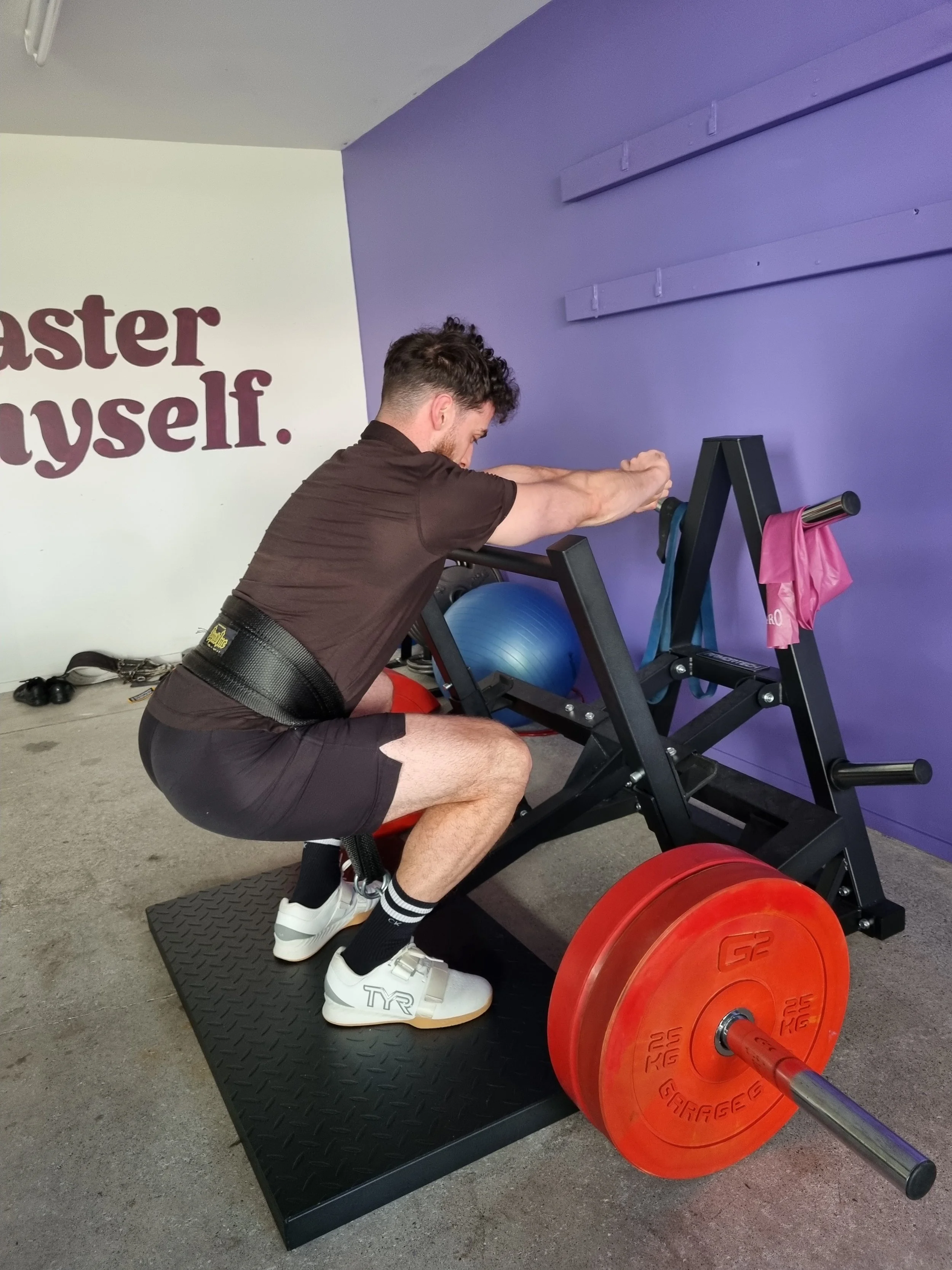 A man wearing a weightlifting belt and sneakers is squatting on a platform with a barbell loaded with large red weight plates, performing a squat exercise in a gym.