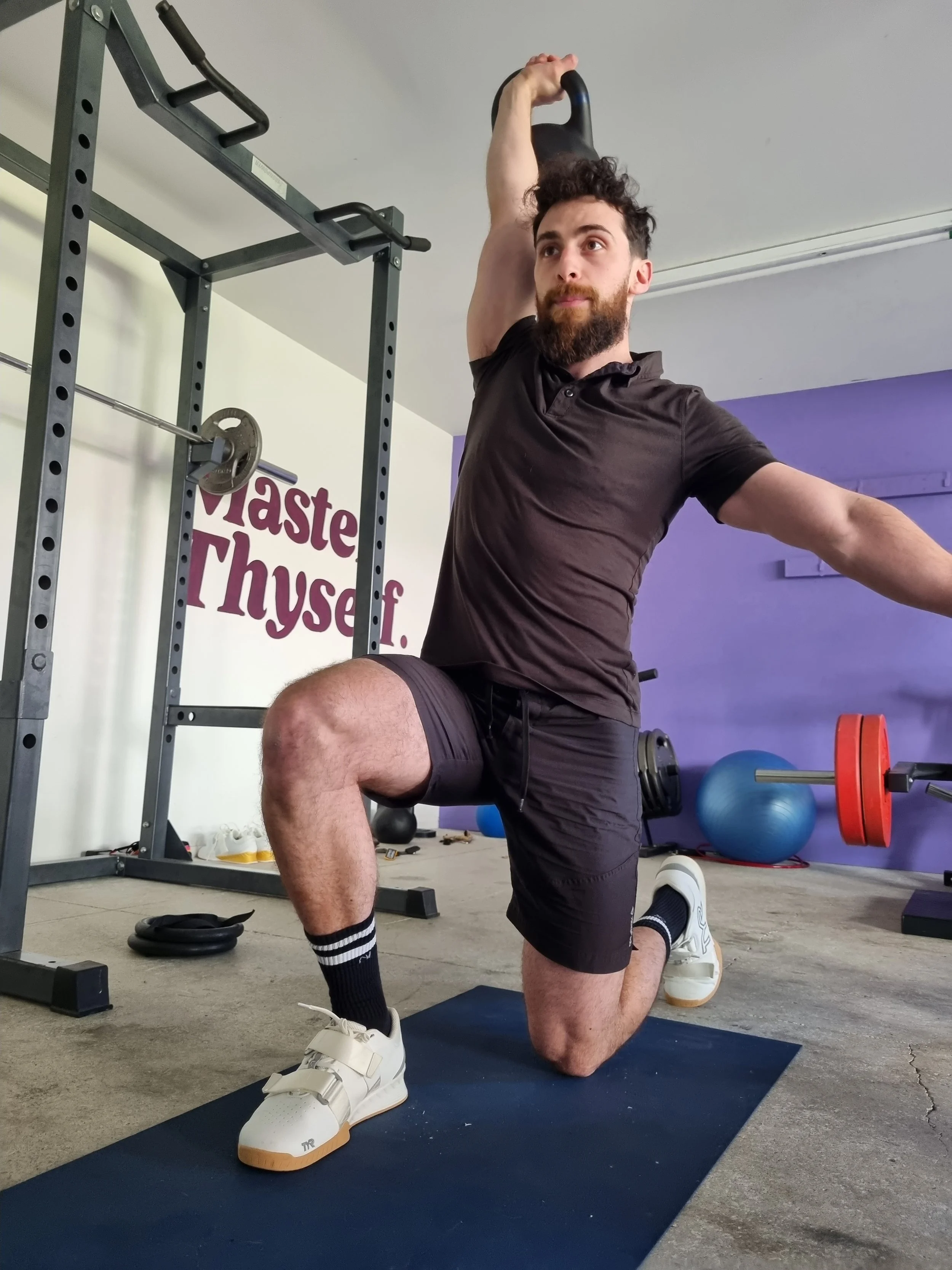 A man with curly hair and a beard in black athletic wear is performing a kettlebell overhead lunge exercise in a gym. He is on a black exercise mat, with gym equipment and a wall with enhances in the background.
