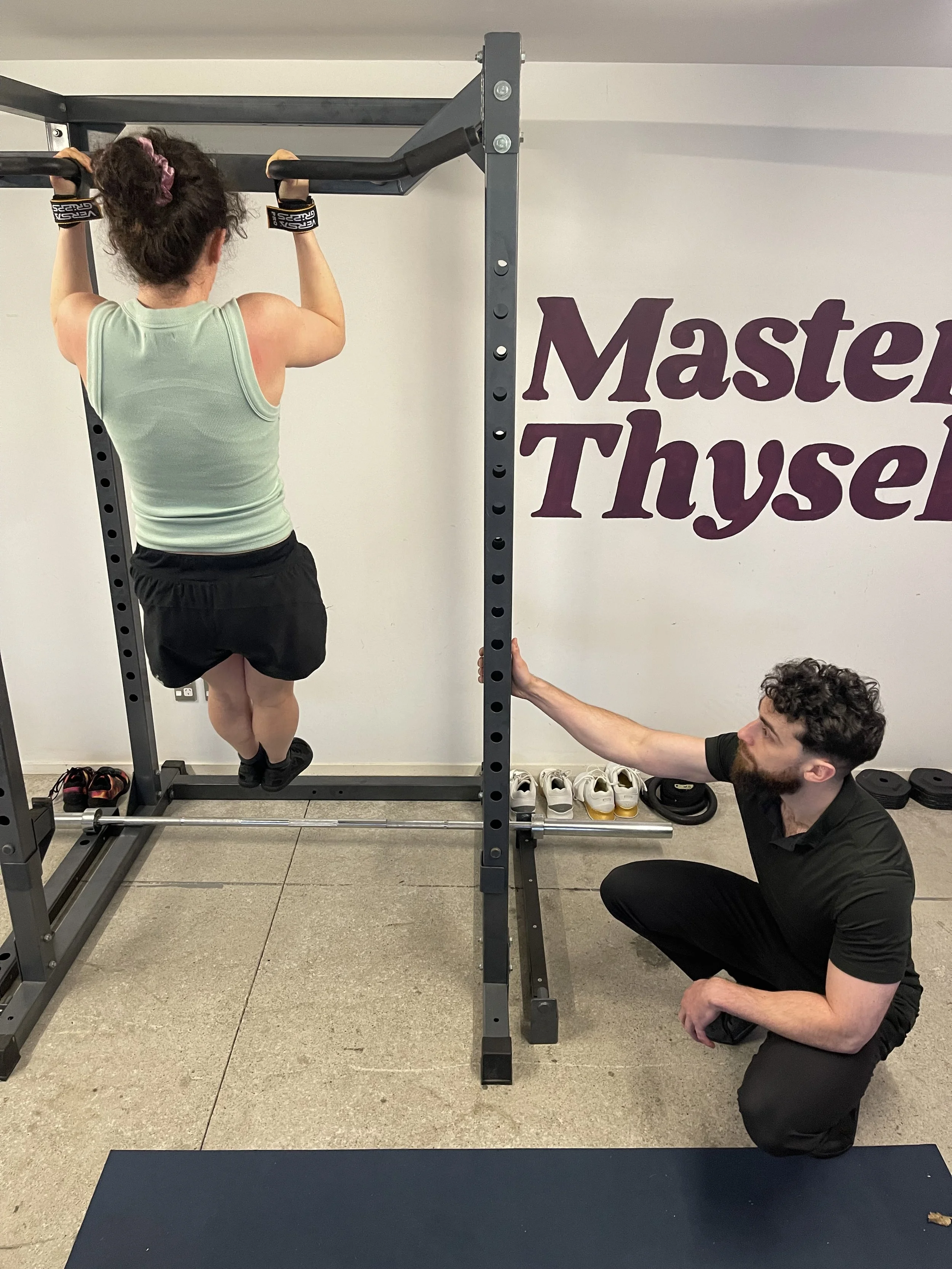 A woman is performing a pull-up on a pull-up bar in a gym, assisted by a trainer who is kneeling on the floor, holding her hand for support. The gym has a sign on the wall that says 'Master Thyself,' with shoes placed on a rack in the background.