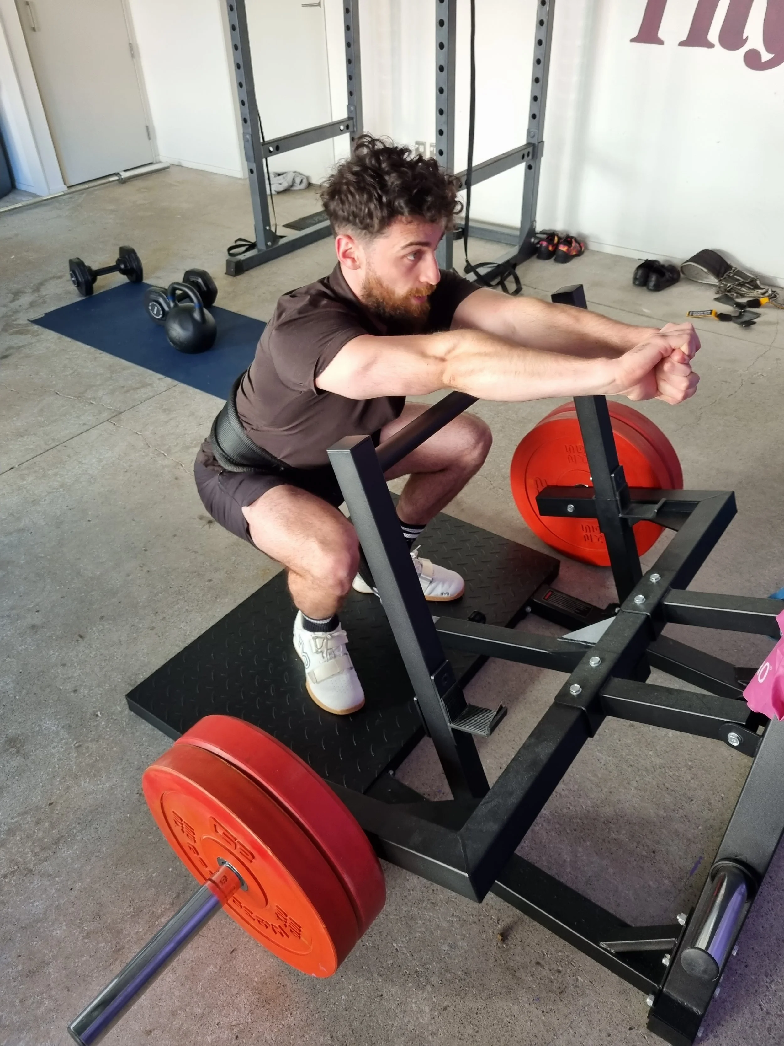 A man with curly hair and a beard, performing a squat exercise on a squat rack with red weight plates in a gym. He is wearing a brown shirt, black shorts, white sneakers, and a weightlifting belt.