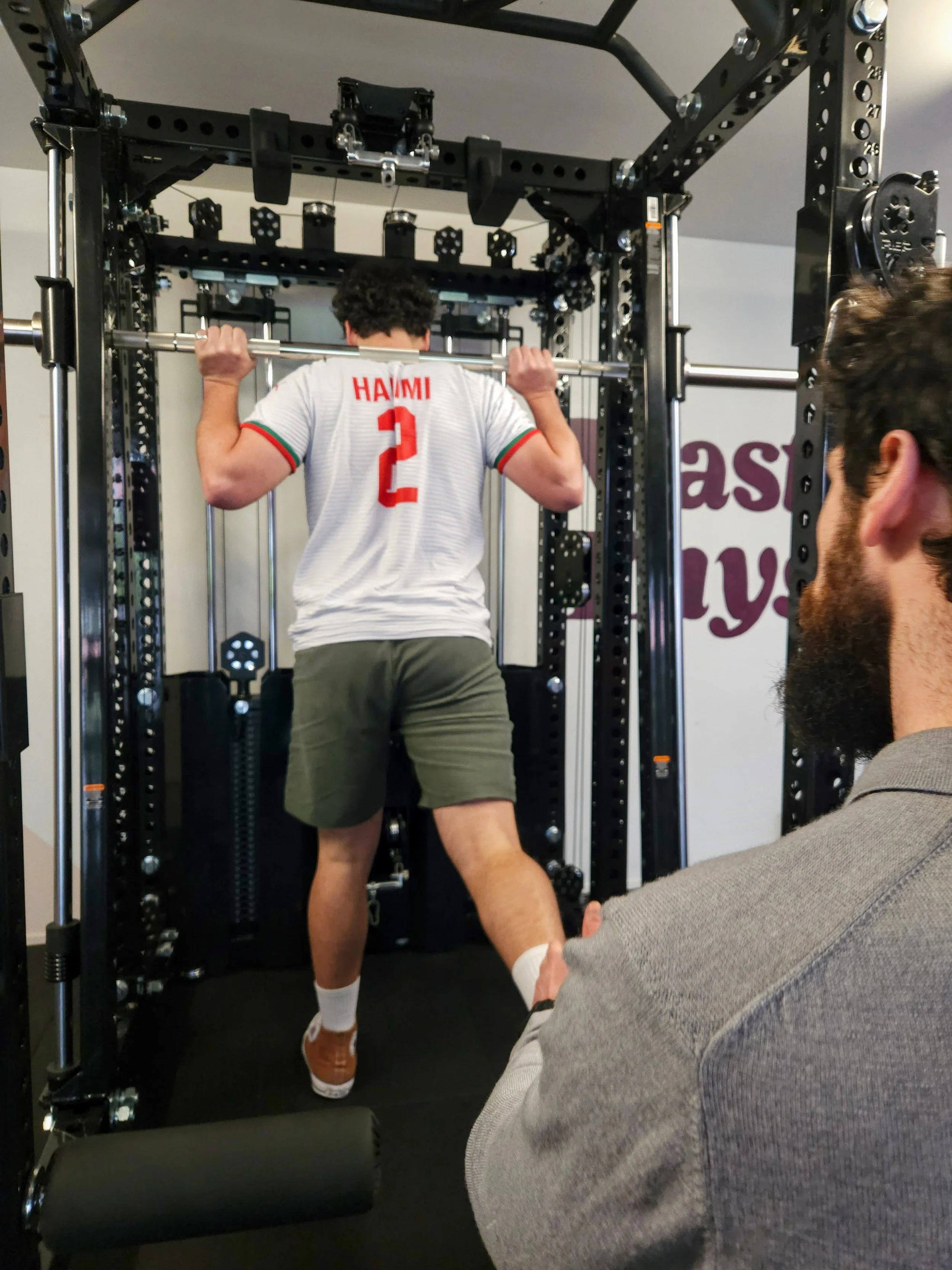A man in athletic clothing preparing to do a Bulgarian split squat with the REP Ares™ 2.0 Smith Machine Rack Attachment, being trained by Julian Etchegoin, Craft Fitness Studio Founder and personal trainer.
