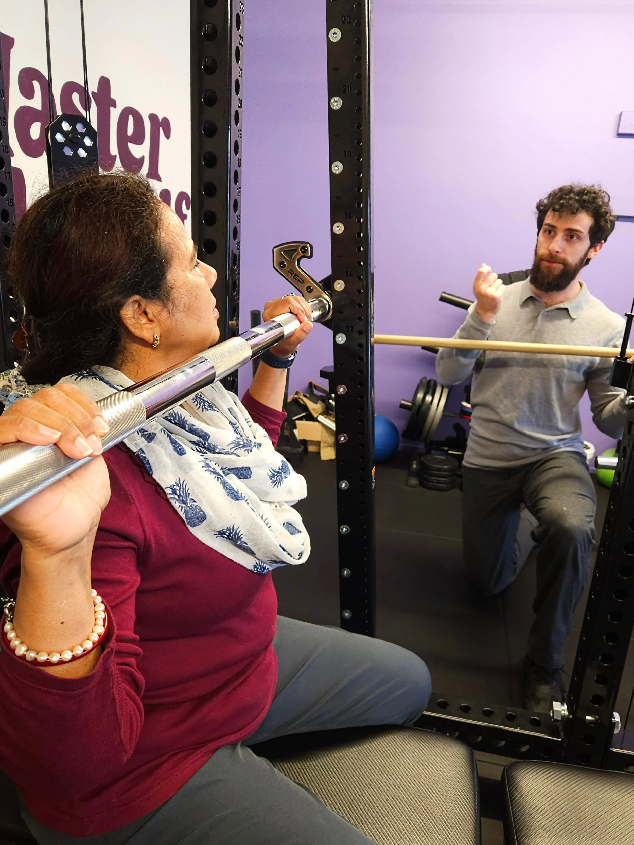 A local Karori woman being instructed through an overhead press exercise using the REP Ares™ 2.0 Smith Machine Rack Attachment. It is placed on her shoulders. Julian, Founder of Craft Fitness Studio is next to her and there are purple walls and worko